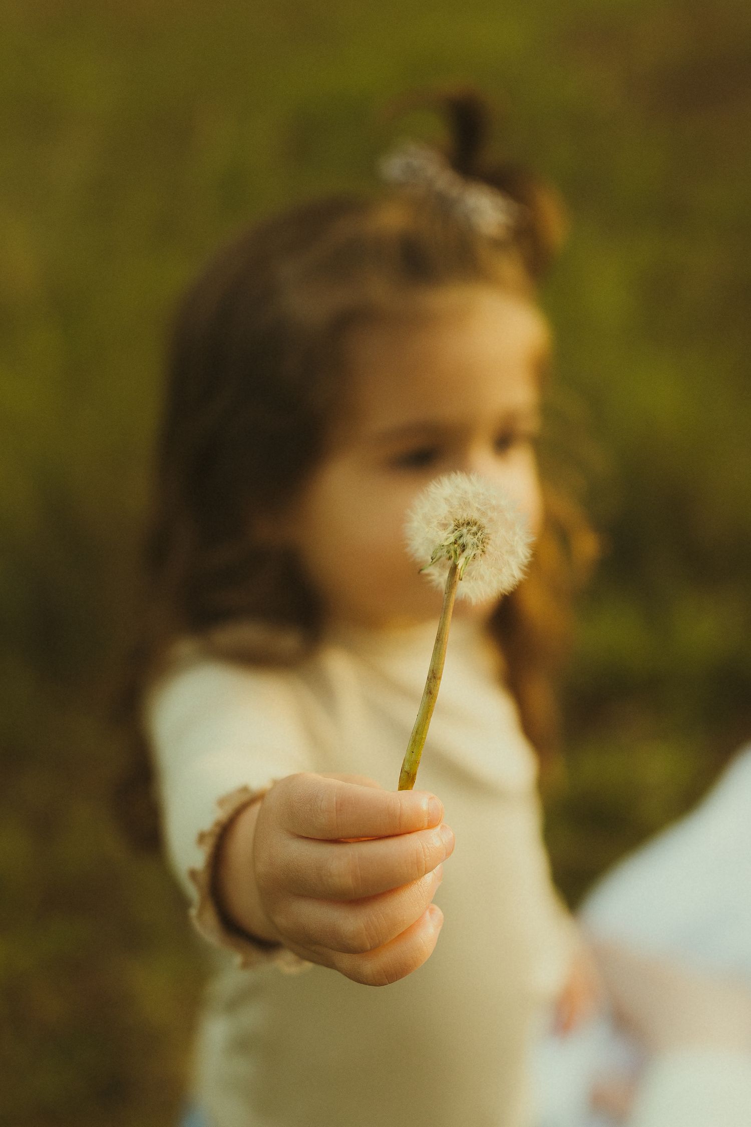 Small hands hold a delicate dandelion against a soft-focused nature background.
