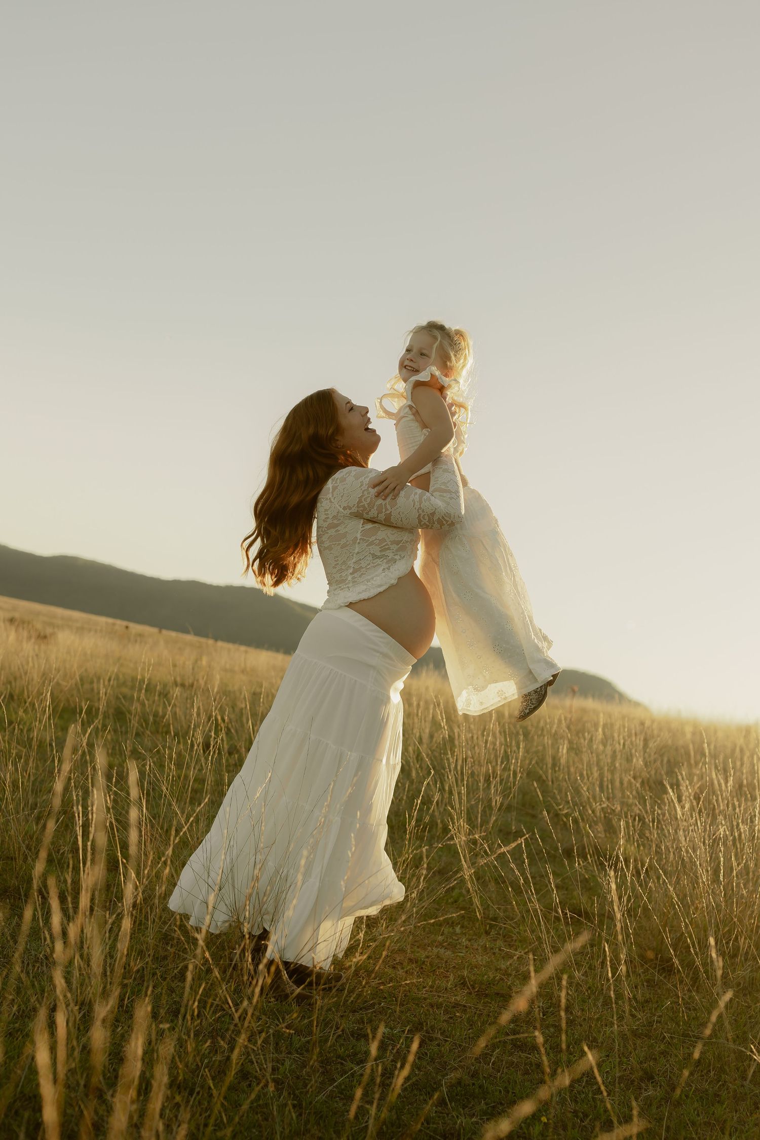 A mother in a white dress lifts her baby in a sunlit golden field at sunset.