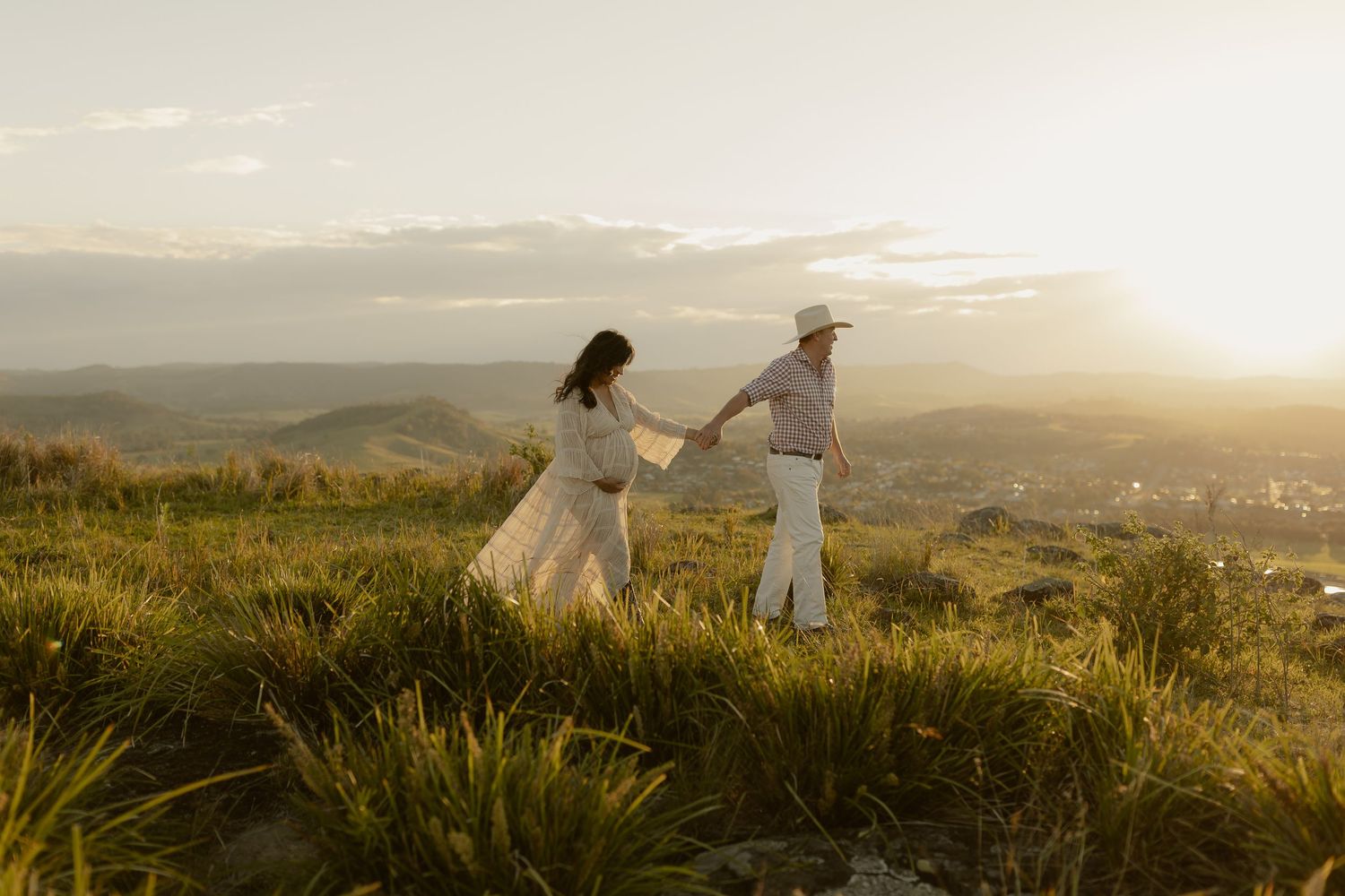 Couple in white attire holding hands at golden sunset on a lush green hilltop with scenic valley views.