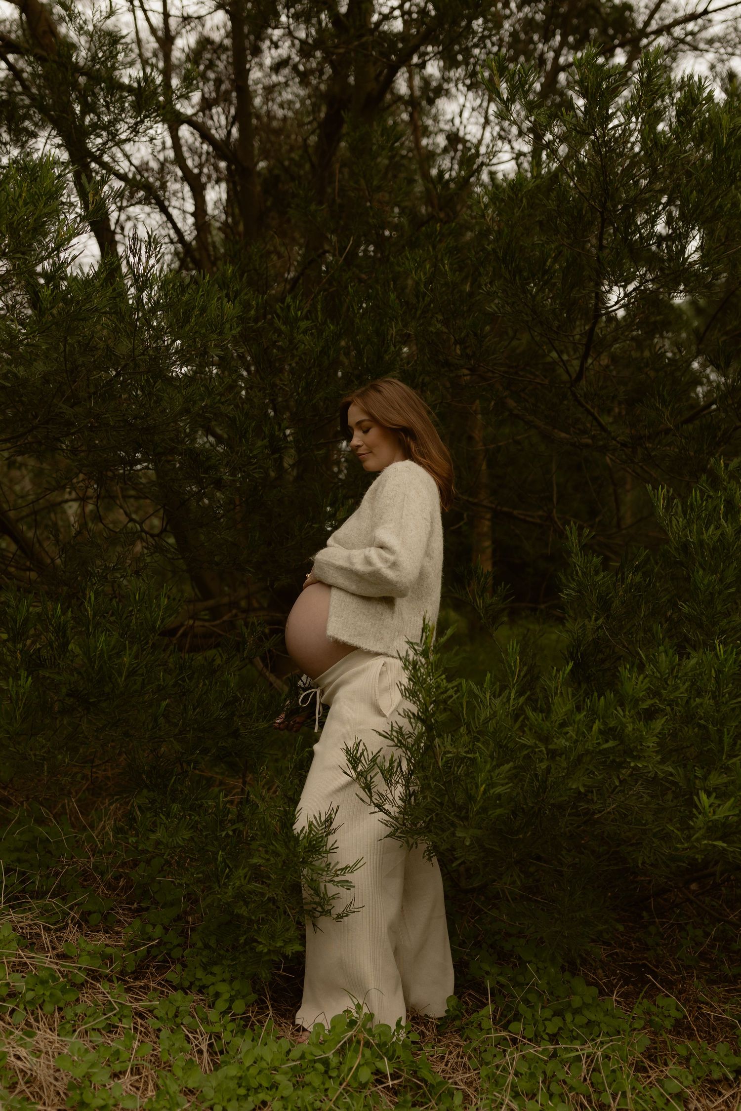 Side profile of pregnant woman in white sweater holding belly surrounded by green shrubs.