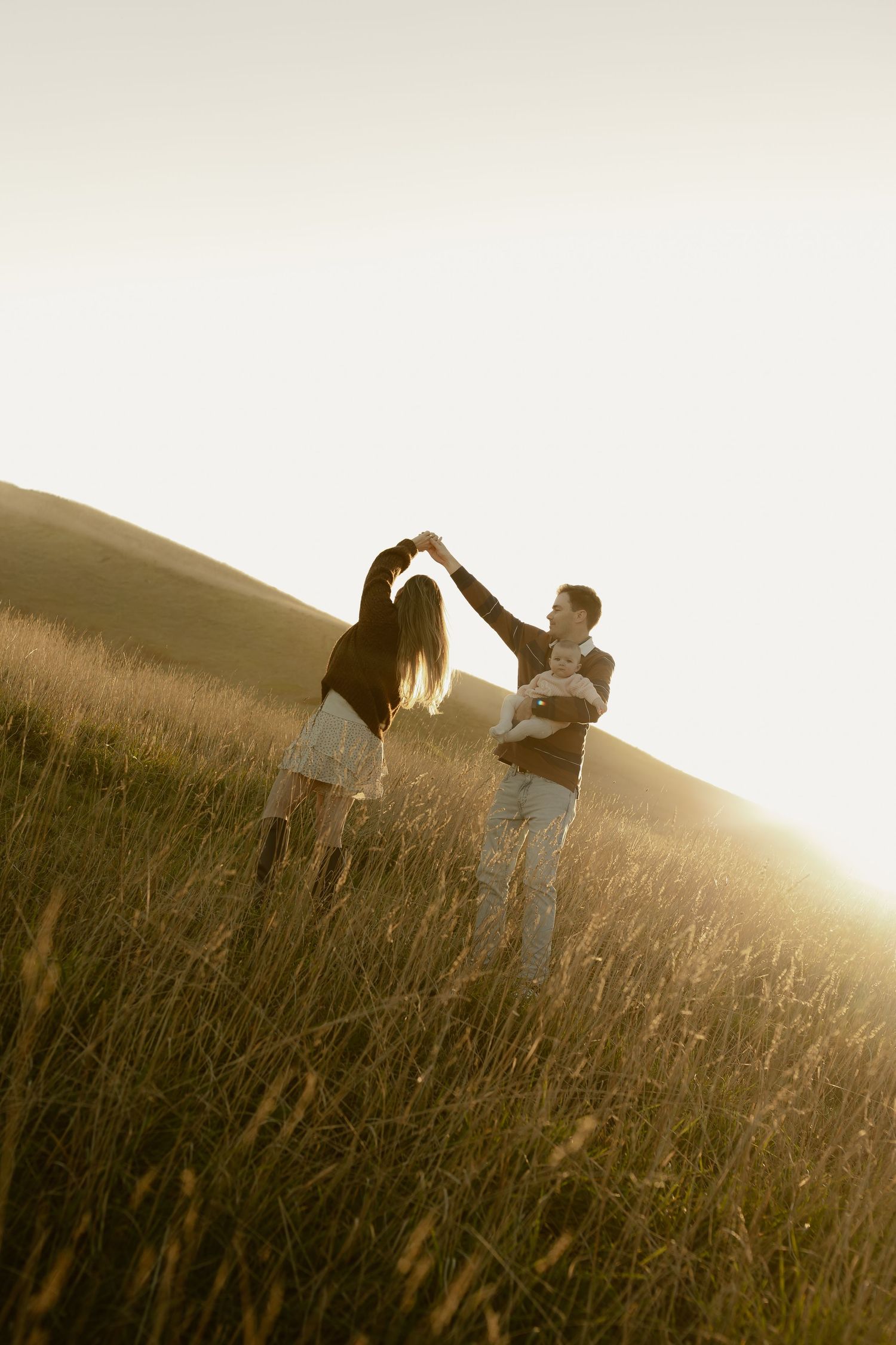 A couple dances in a sunlit golden field while the man holds their baby at sunset.