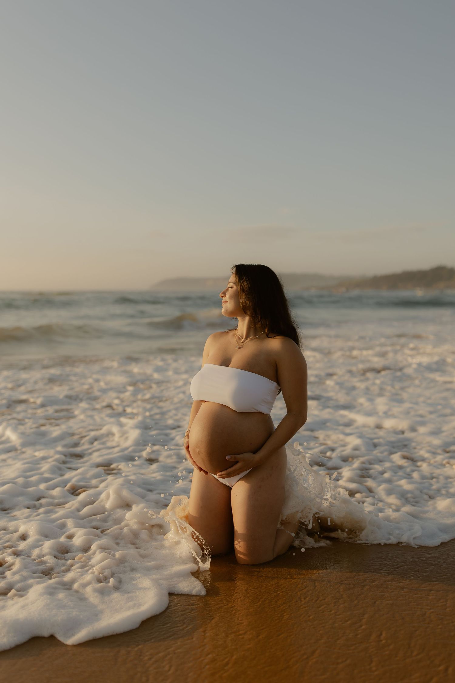 A pregnant woman in white swimwear sits peacefully by the ocean shore as waves roll in at sunset.