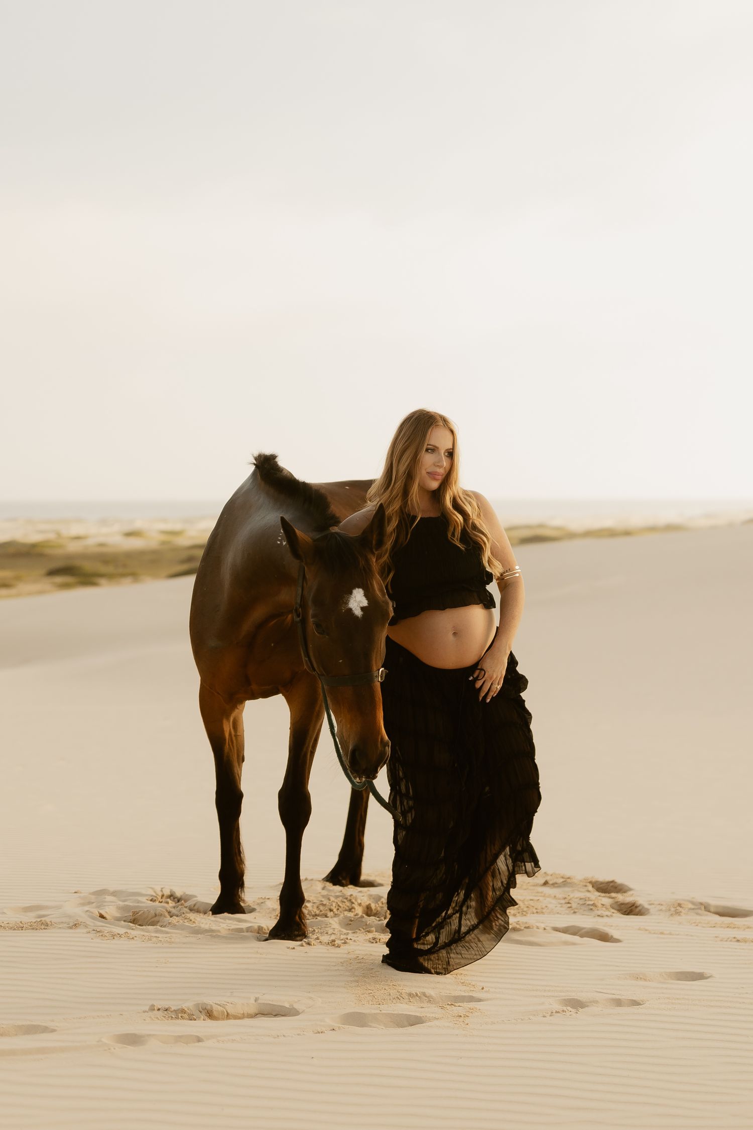 A woman in a black dress poses elegantly next to a dark horse on sandy dunes in warm lighting.