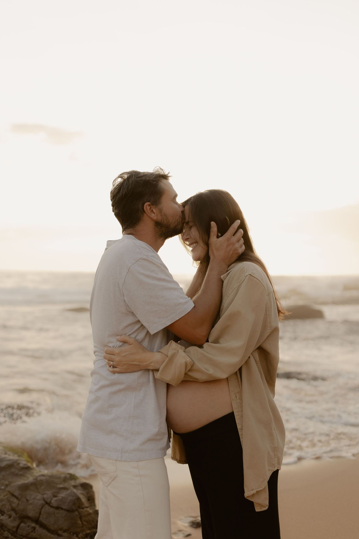 A romantic moment between two people embracing on a beach during golden hour with ocean waves in the background.