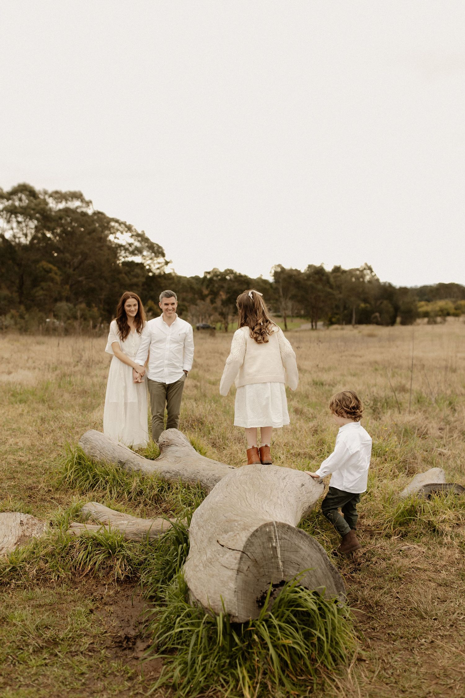 A romantic couple poses playfully around a large rock formation in a rustic outdoor field setting.