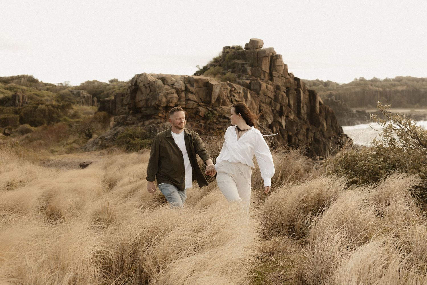 A couple holding hands walks through tall grass on a rocky coastal cliff during golden hour.