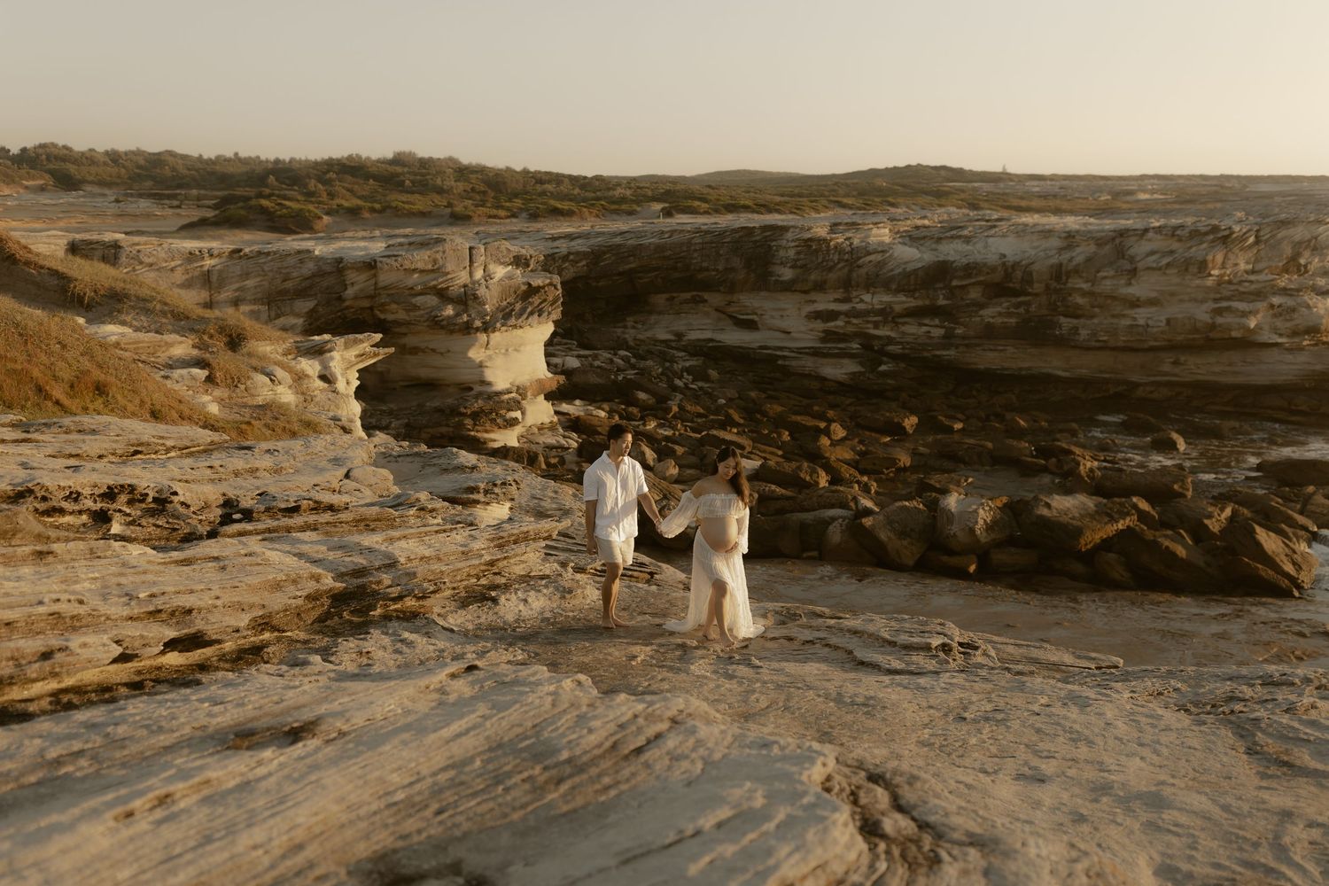 Scenic coastal landscape showing rocky cliffs and two distant figures walking at sunset.
