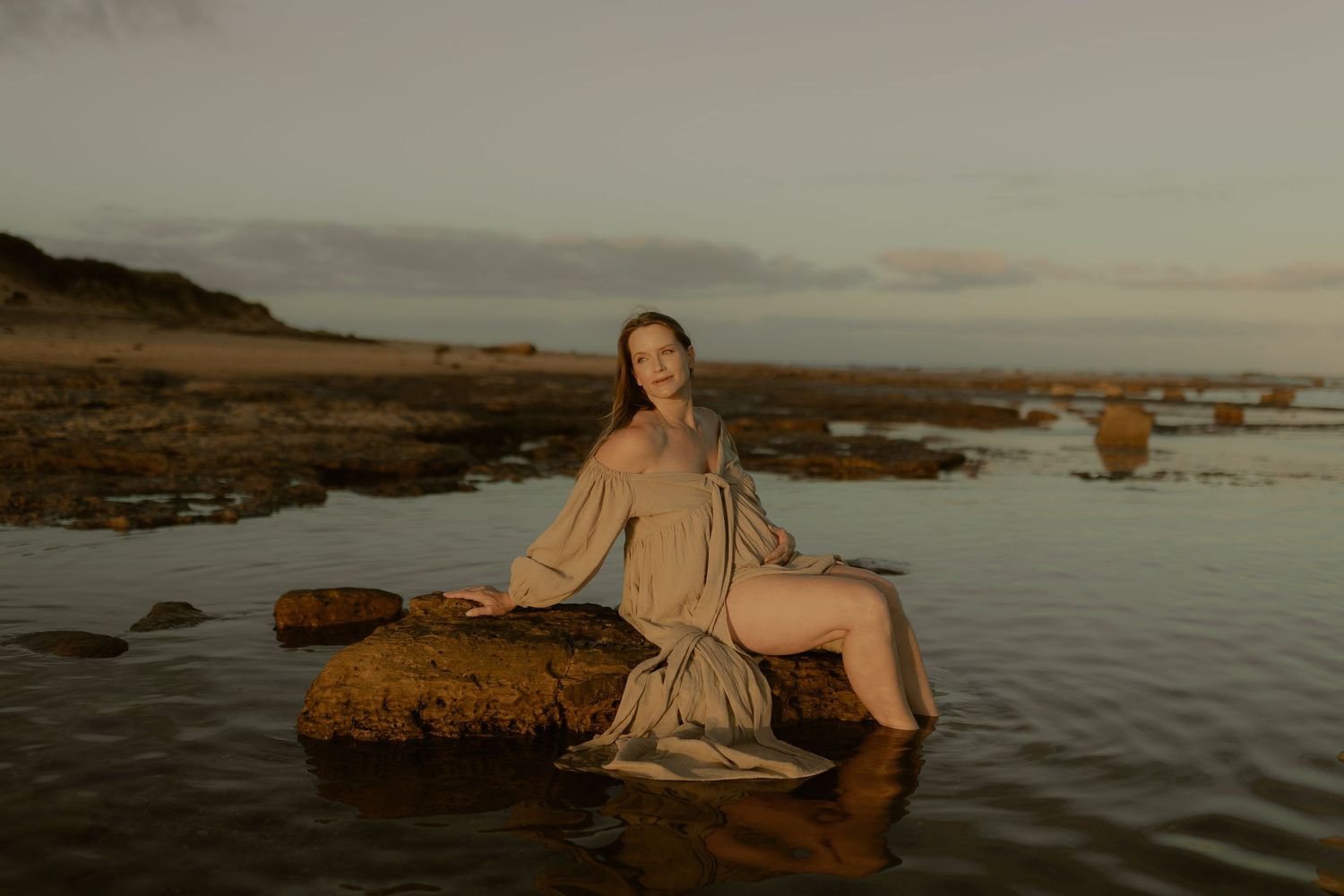 Dramatic coastal scene with figures sitting on rocky shore at golden hour as waves lap against the coastline.