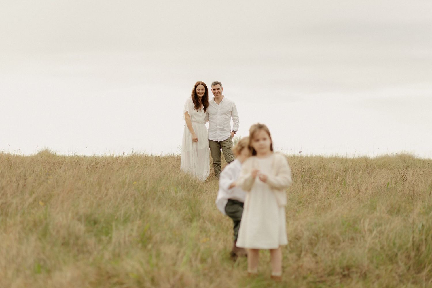Three figures in light colored dresses pose together in a serene field at sunset.