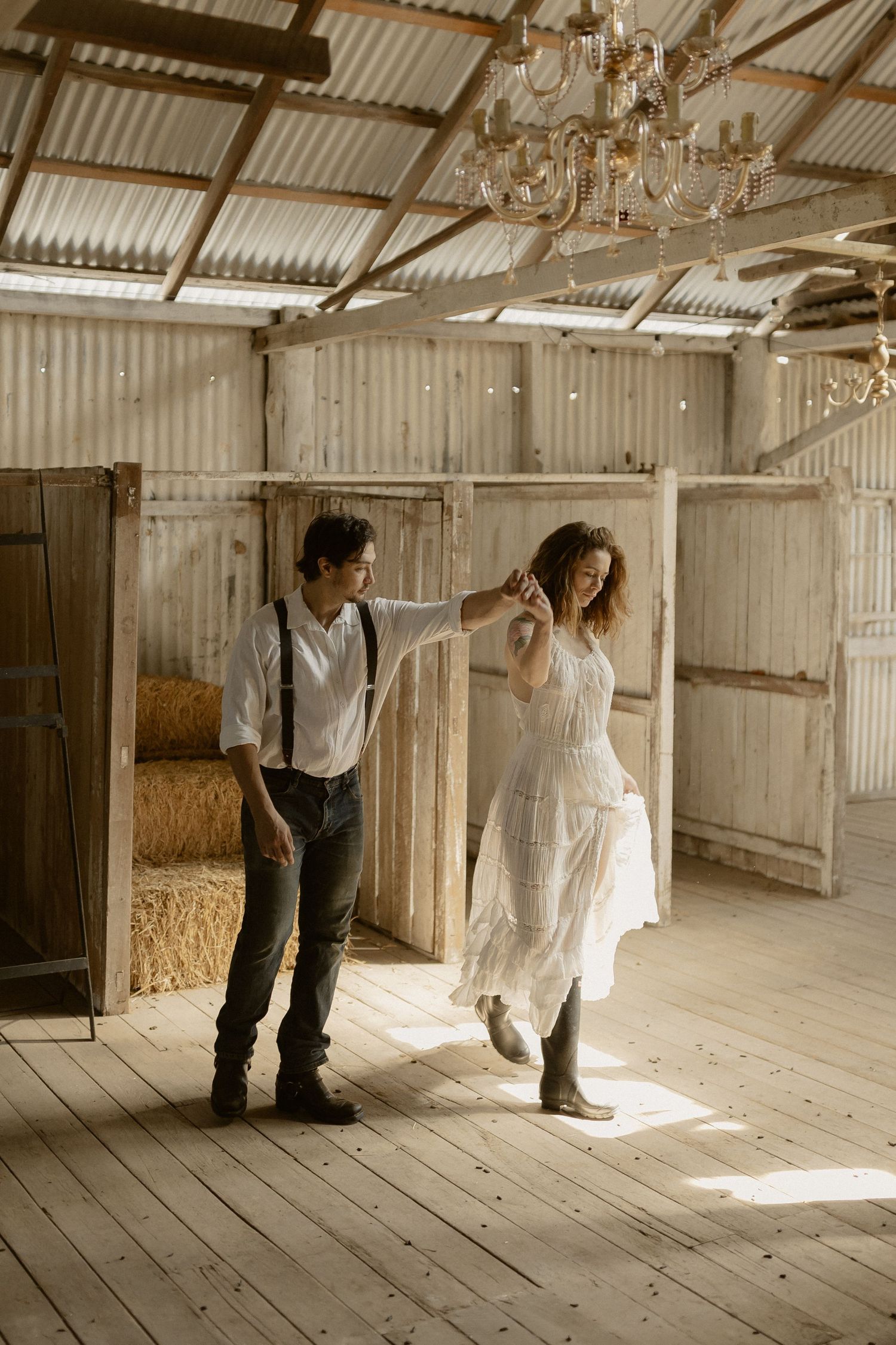 Two figures stand in a sunlit rustic barn with wooden floors and hay bales visible in the background.