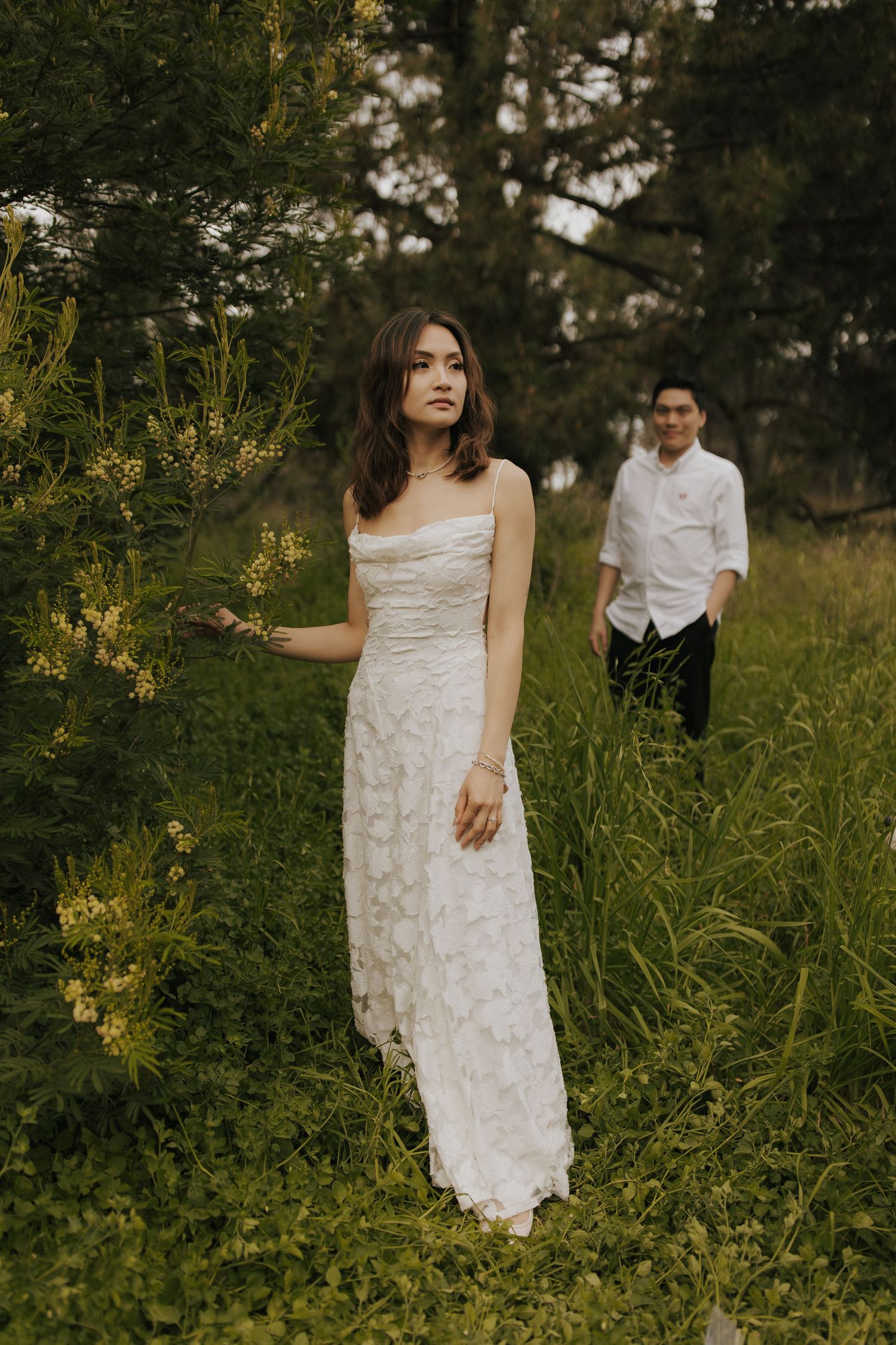 A couple poses together in a grassy field wearing white clothing during a romantic photoshoot.