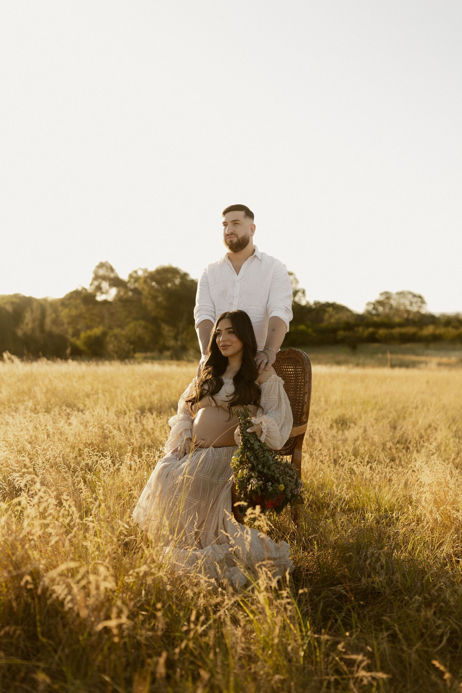 Sunset maternity session with couple in white standing among tall golden grasses.