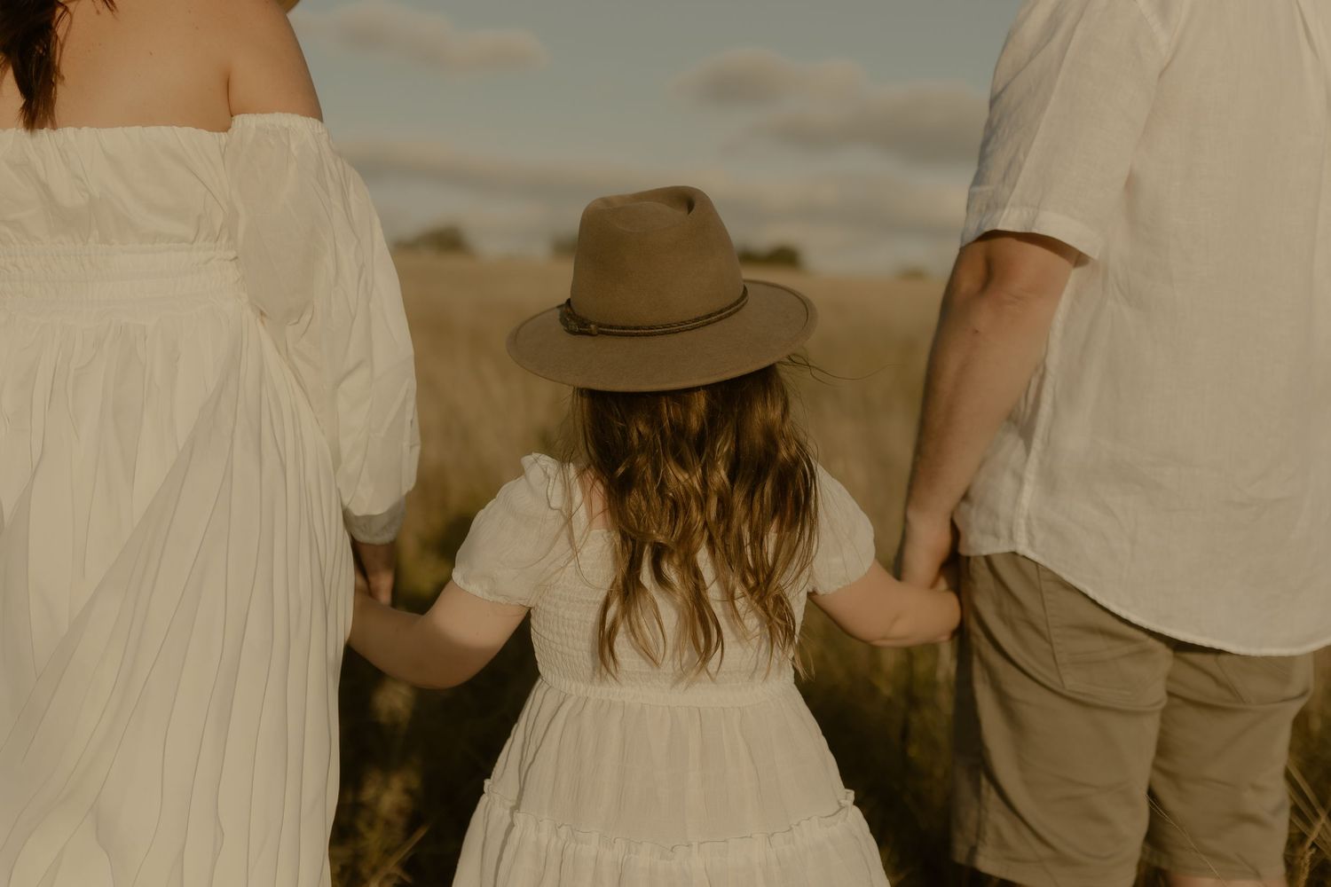 A child in a brown hat and white dress holds hands with their parents on a beach at sunset.