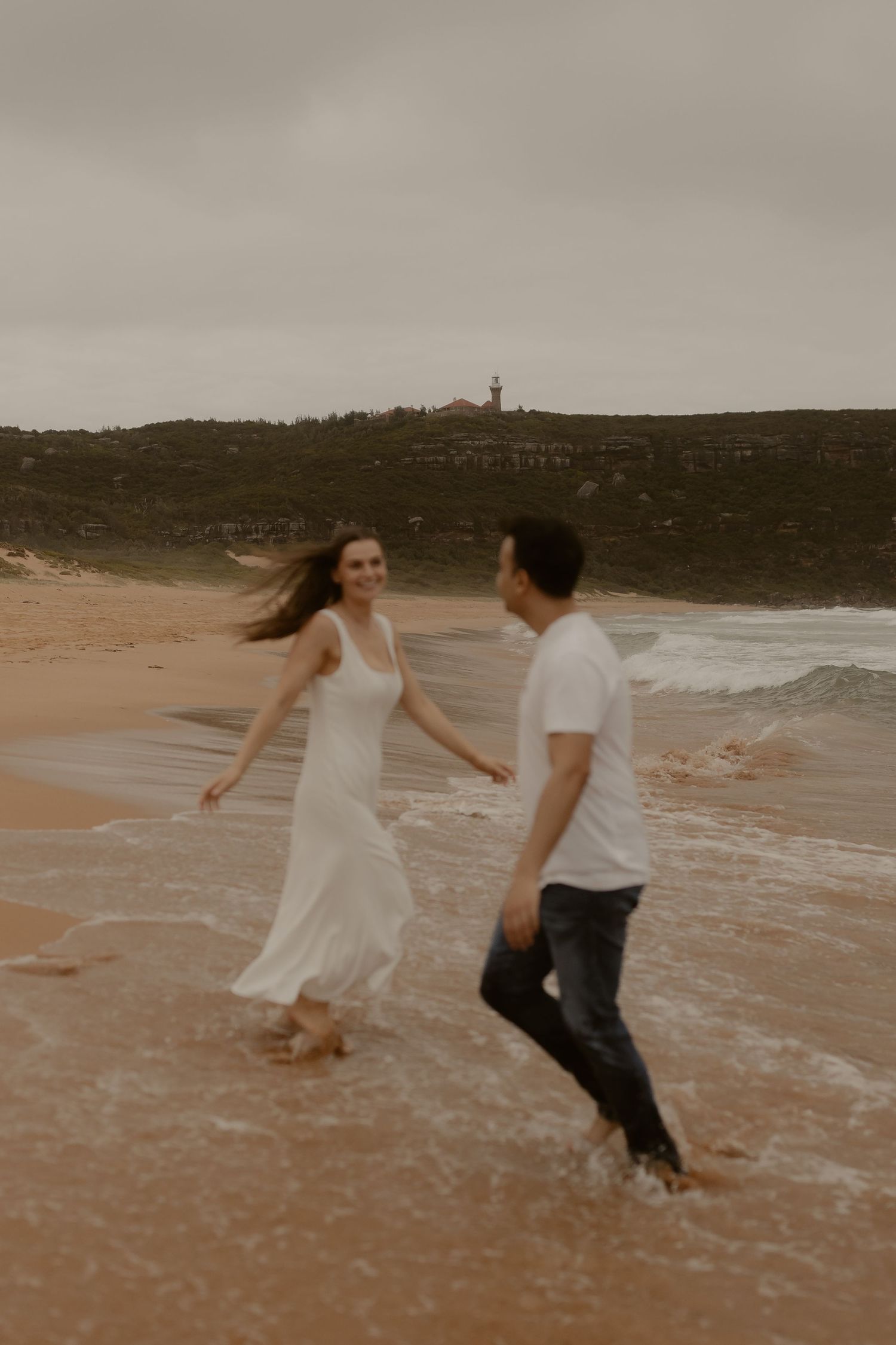 Two people dance playfully in the shallow waves on a cloudy beach day.