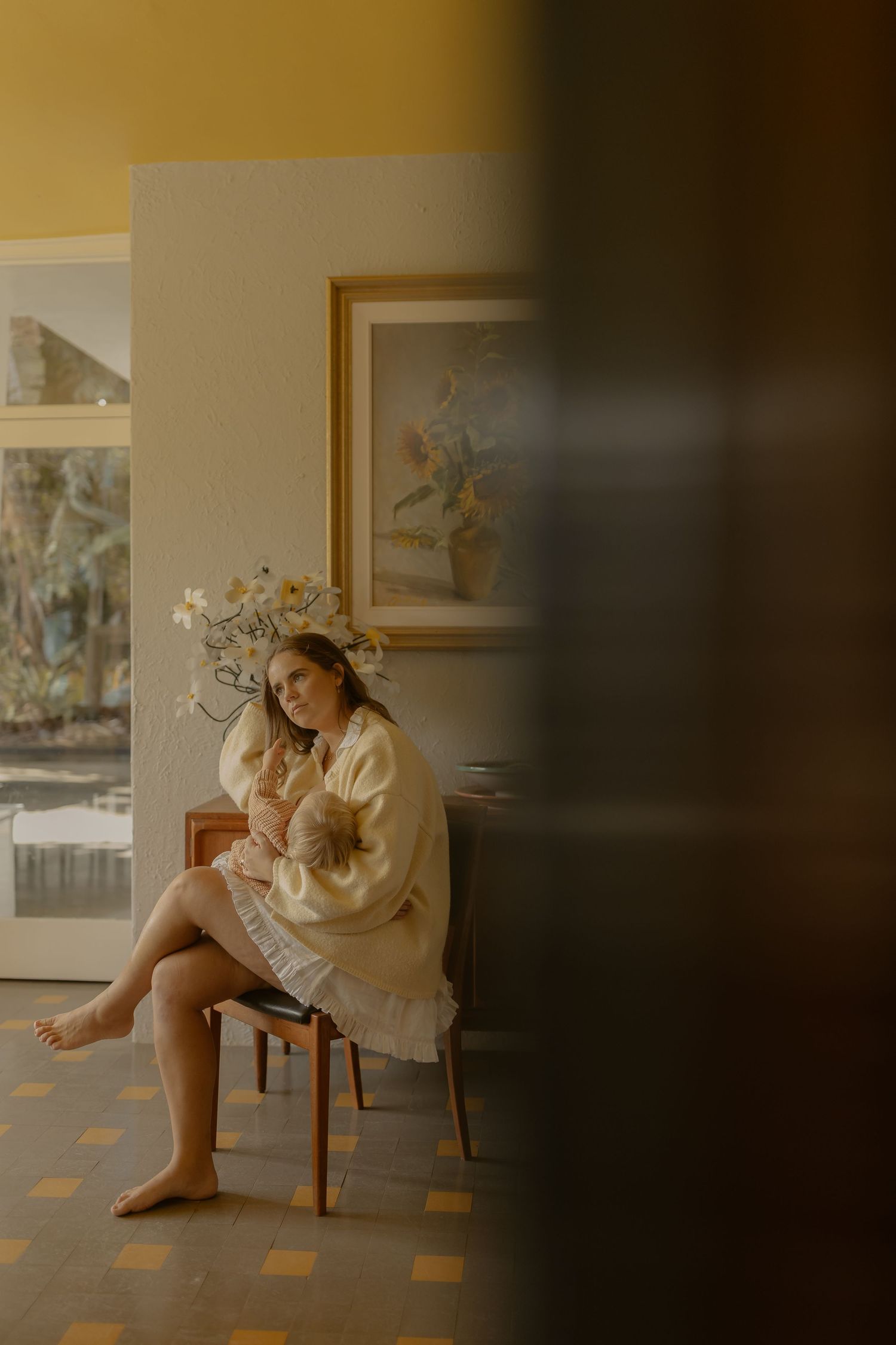 A barefoot woman in a cream cardigan sits on a chair beside white flowers with a framed floral painting on the wall.