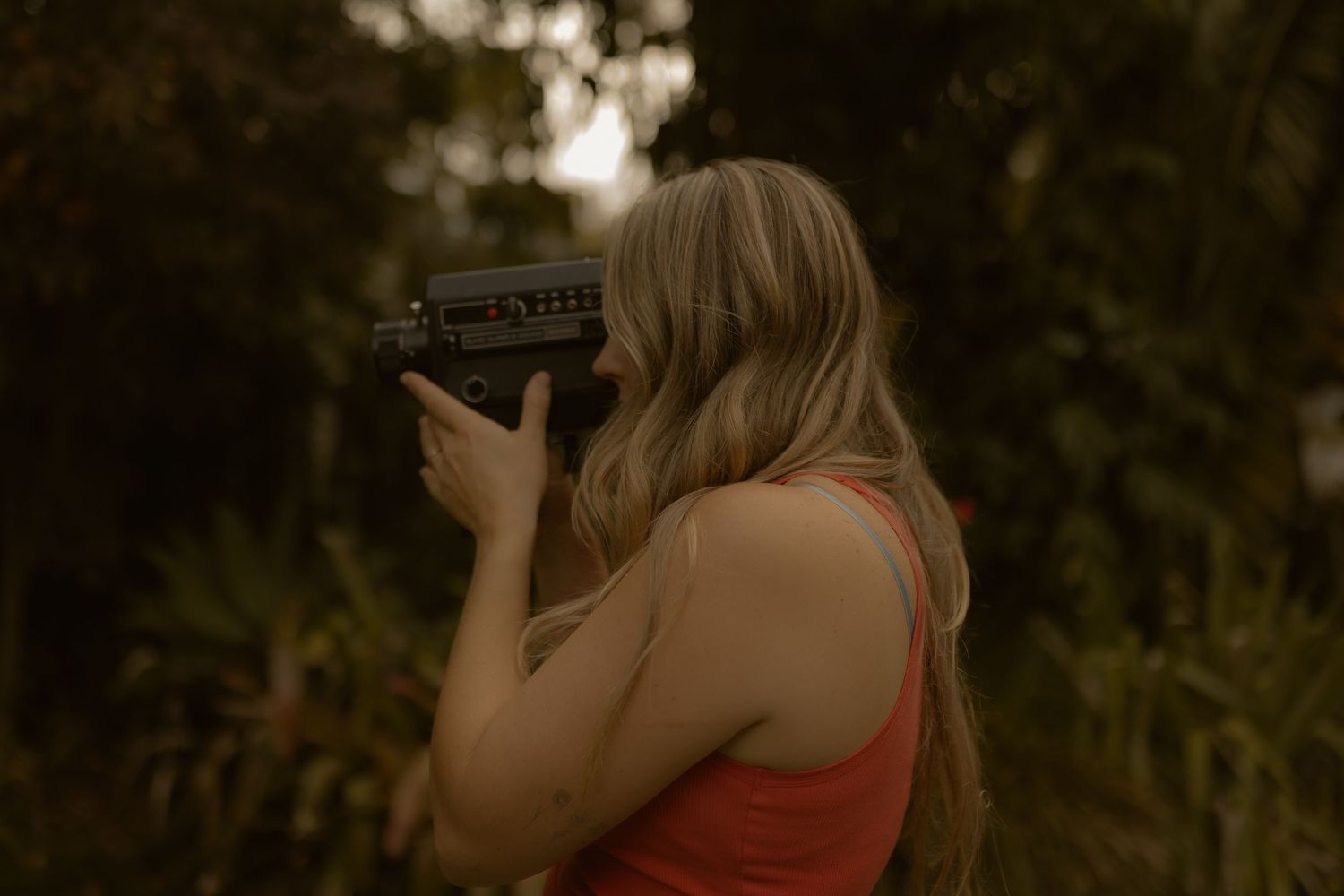 Woman with blonde hair in red top aiming a camera outdoors among lush green trees in warm light.