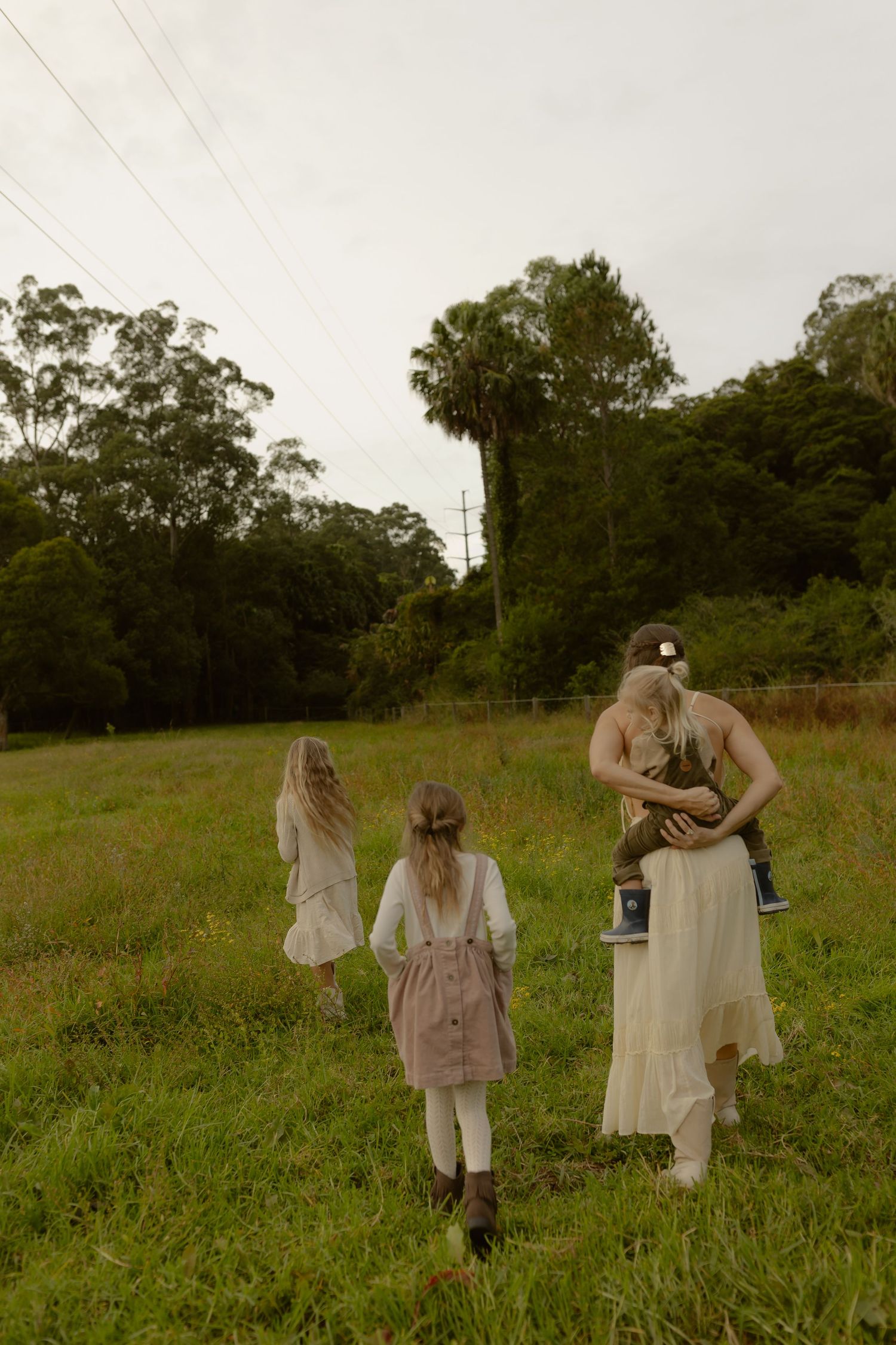 A woman in a cream dress carries a baby while two girls walk ahead through a green grassy field.