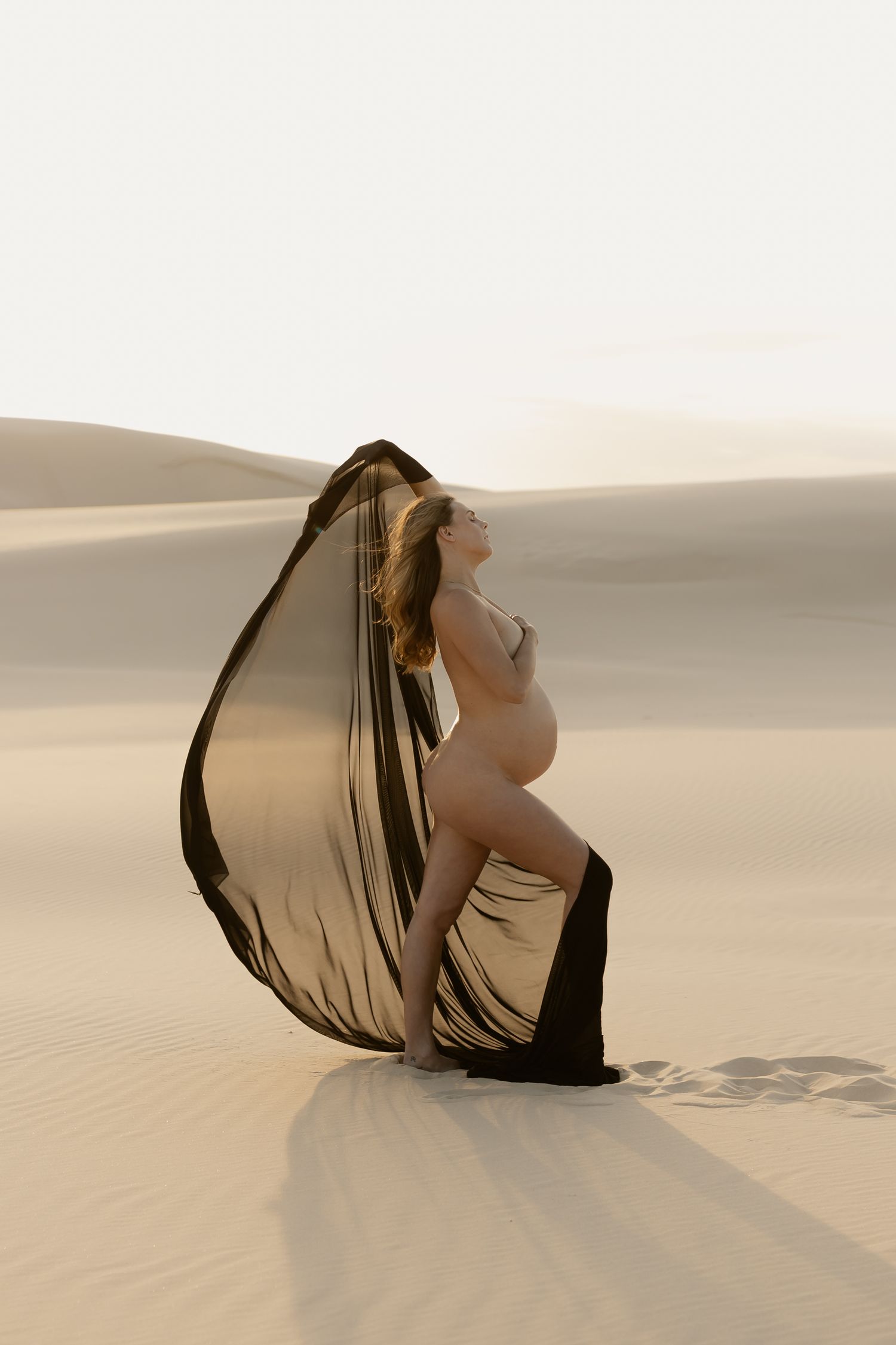 Pregnant woman draped in flowing dark fabric poses gracefully on golden sand dunes in a serene desert maternity photo.