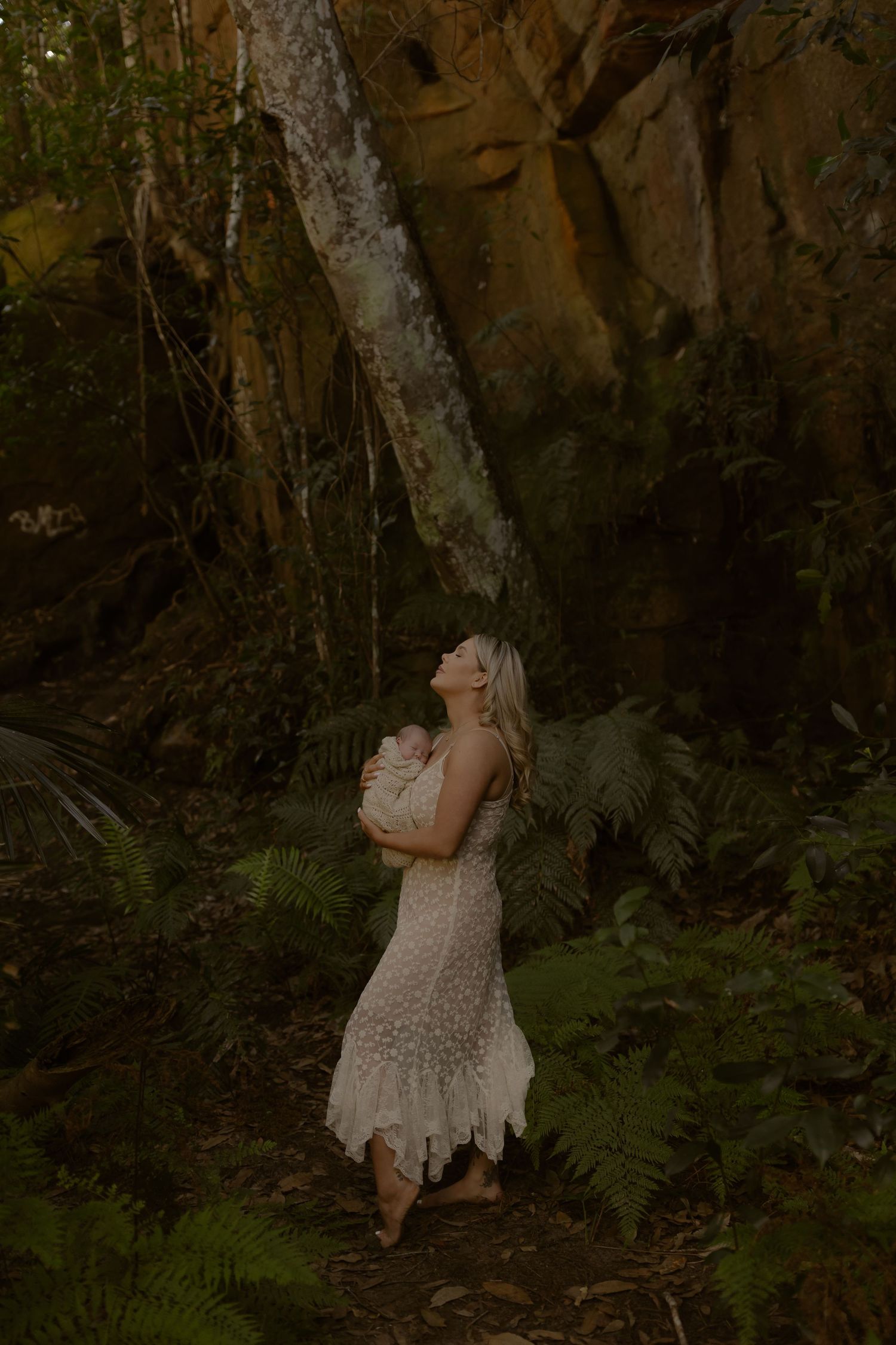 A blonde woman in a white lace dress holds a baby while gazing upward in a lush forest setting.