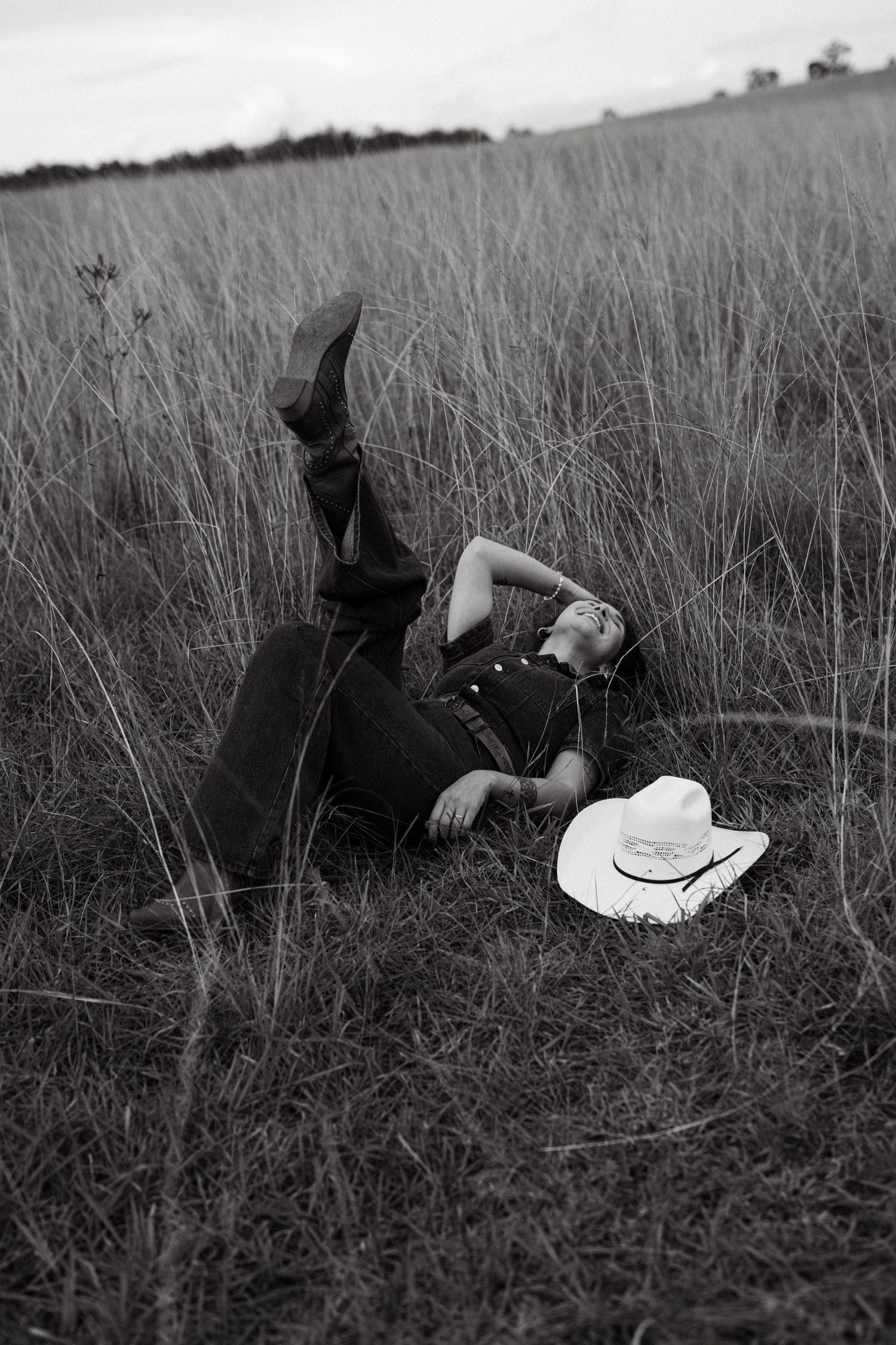 Black and white photo of a cowboy lying in tall grass field with white hat beside them, legs raised, rural landscape.