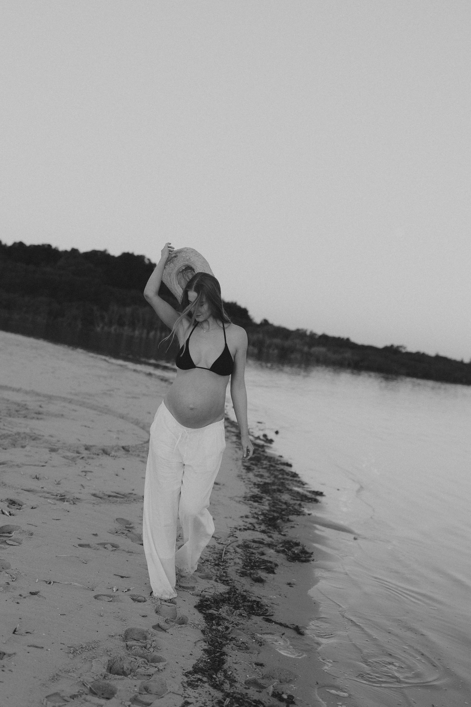 Black and white photo of pregnant woman in bikini and white pants tossing hat on a serene beach at sunset.