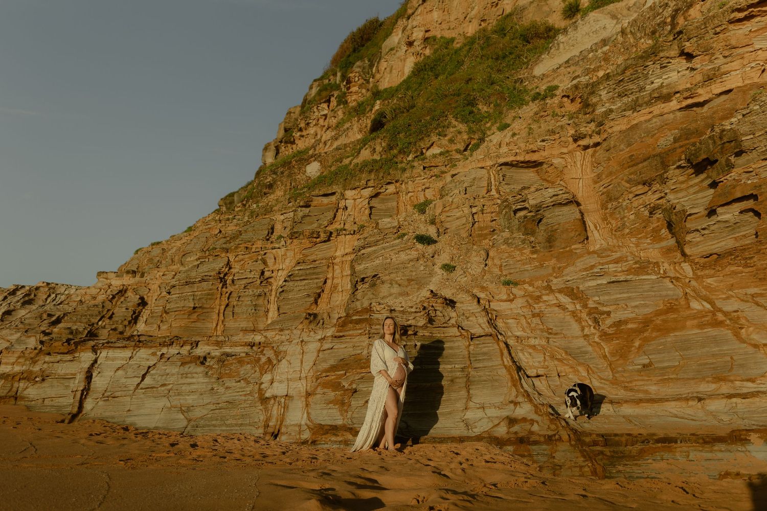 A couple stands against towering layered sandstone cliffs on a sandy beach, dressed in elegant white attire.