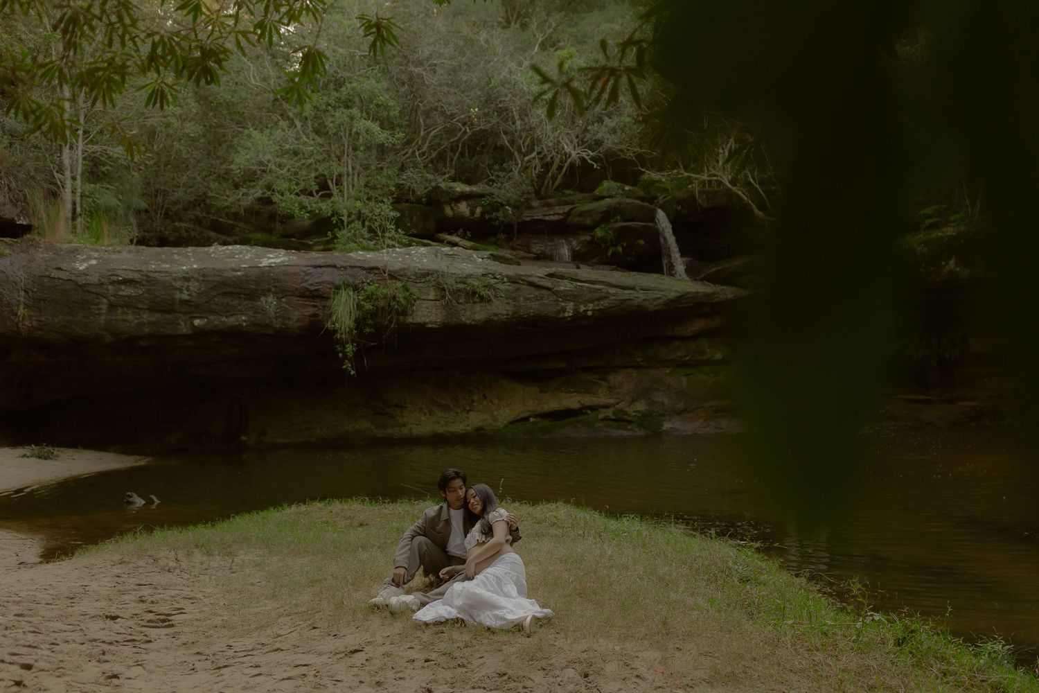 Couple sitting together on sandy shore beside a tranquil green river surrounded by lush forest and sandstone rocks.