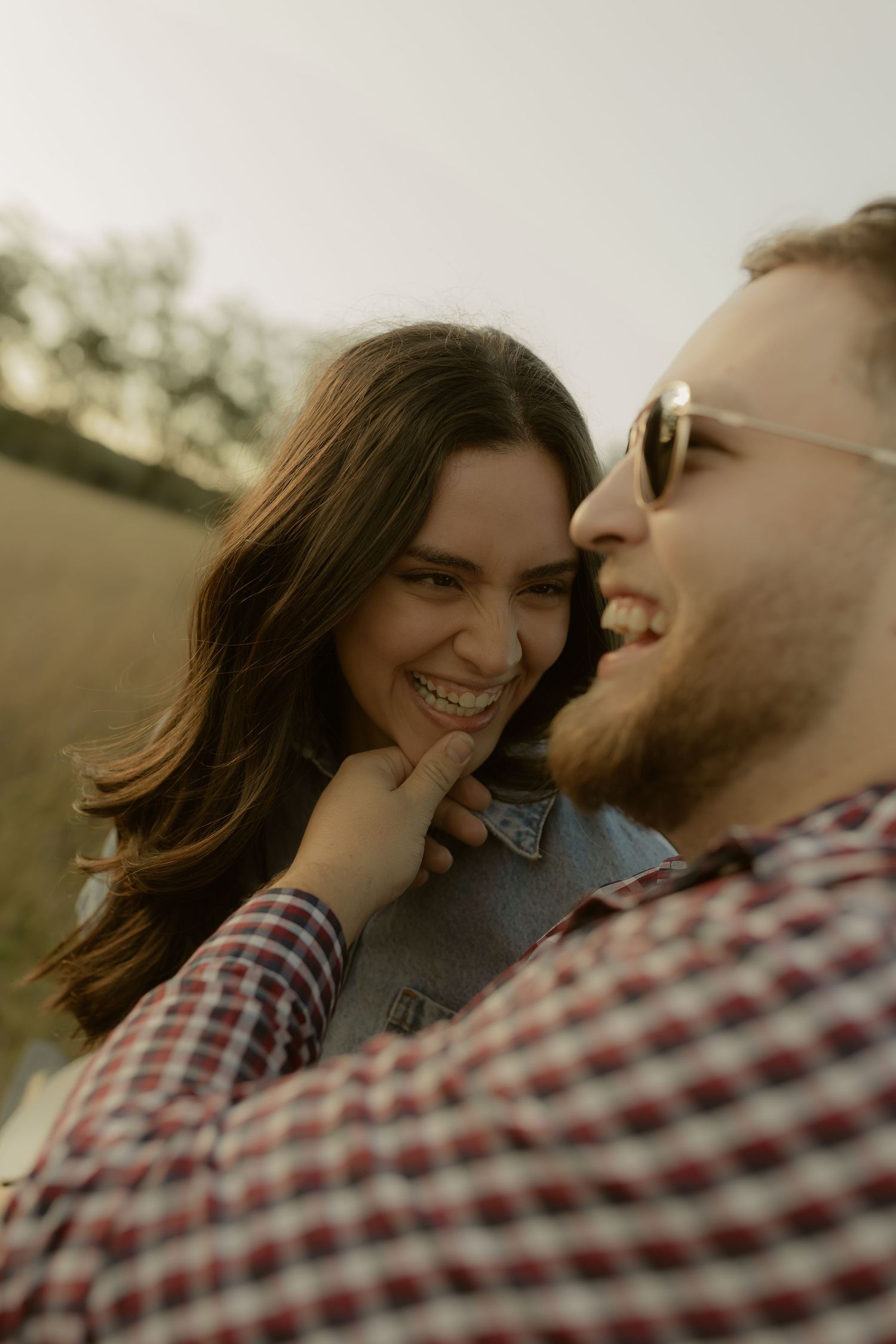 A laughing couple shares a joyful moment outdoors, with the man in sunglasses and plaid shirt holding the smiling woman's chin.