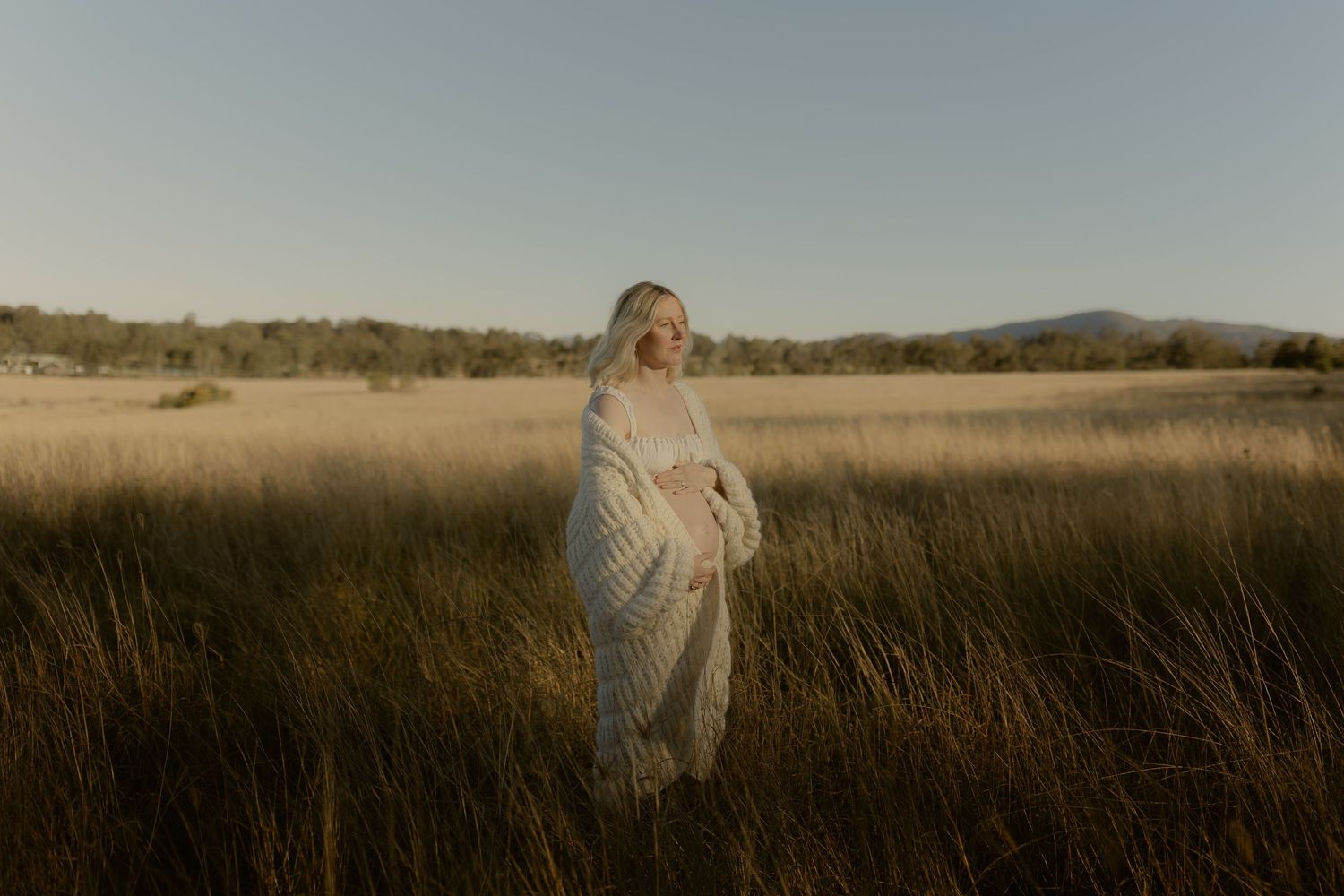 A woman in a flowing white dress stands in a golden field at sunset with mountains in the background.