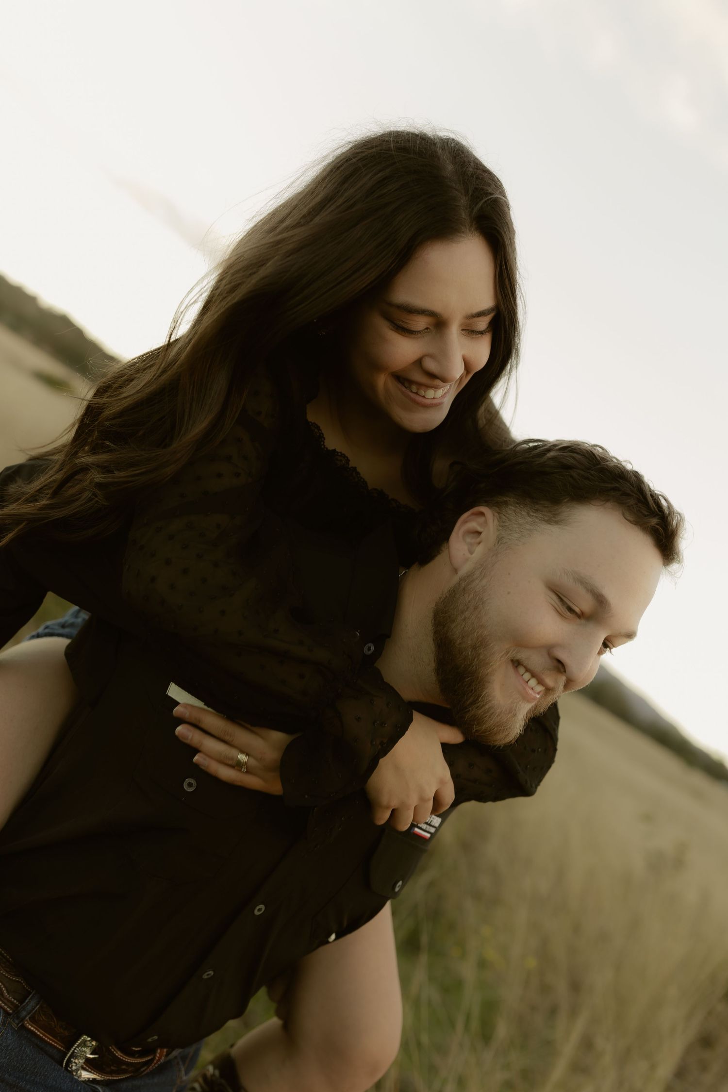 Happy couple in matching black outfits laughing together as woman rides piggyback in grass field.