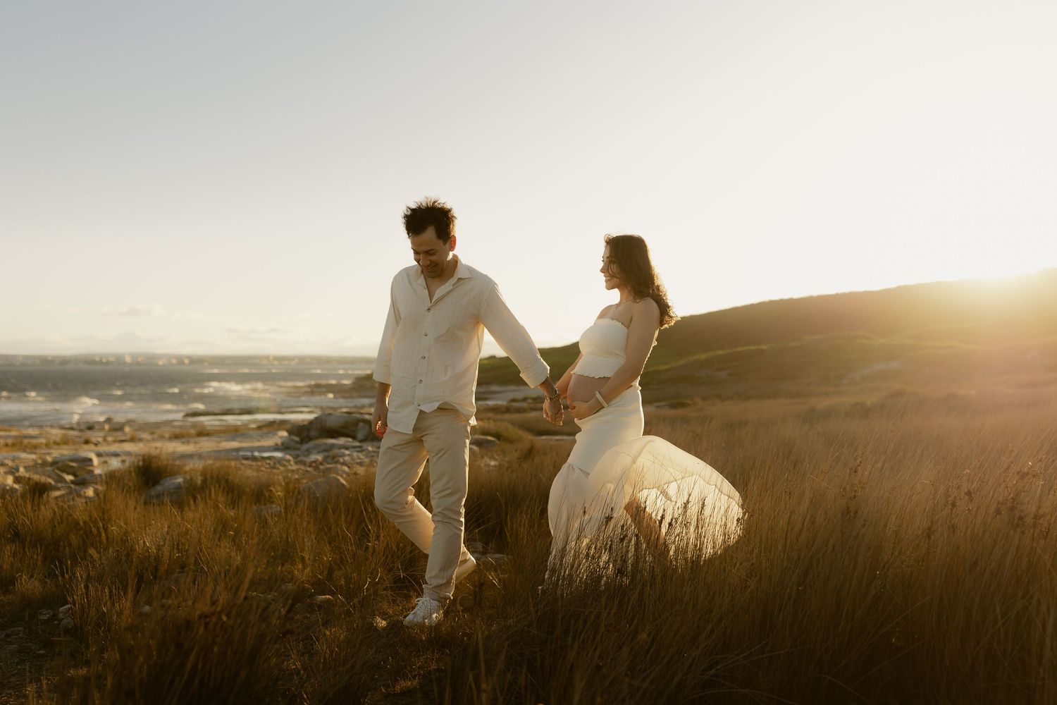Couple in white wedding attire holding hands walking on coastal hillside at golden hour.