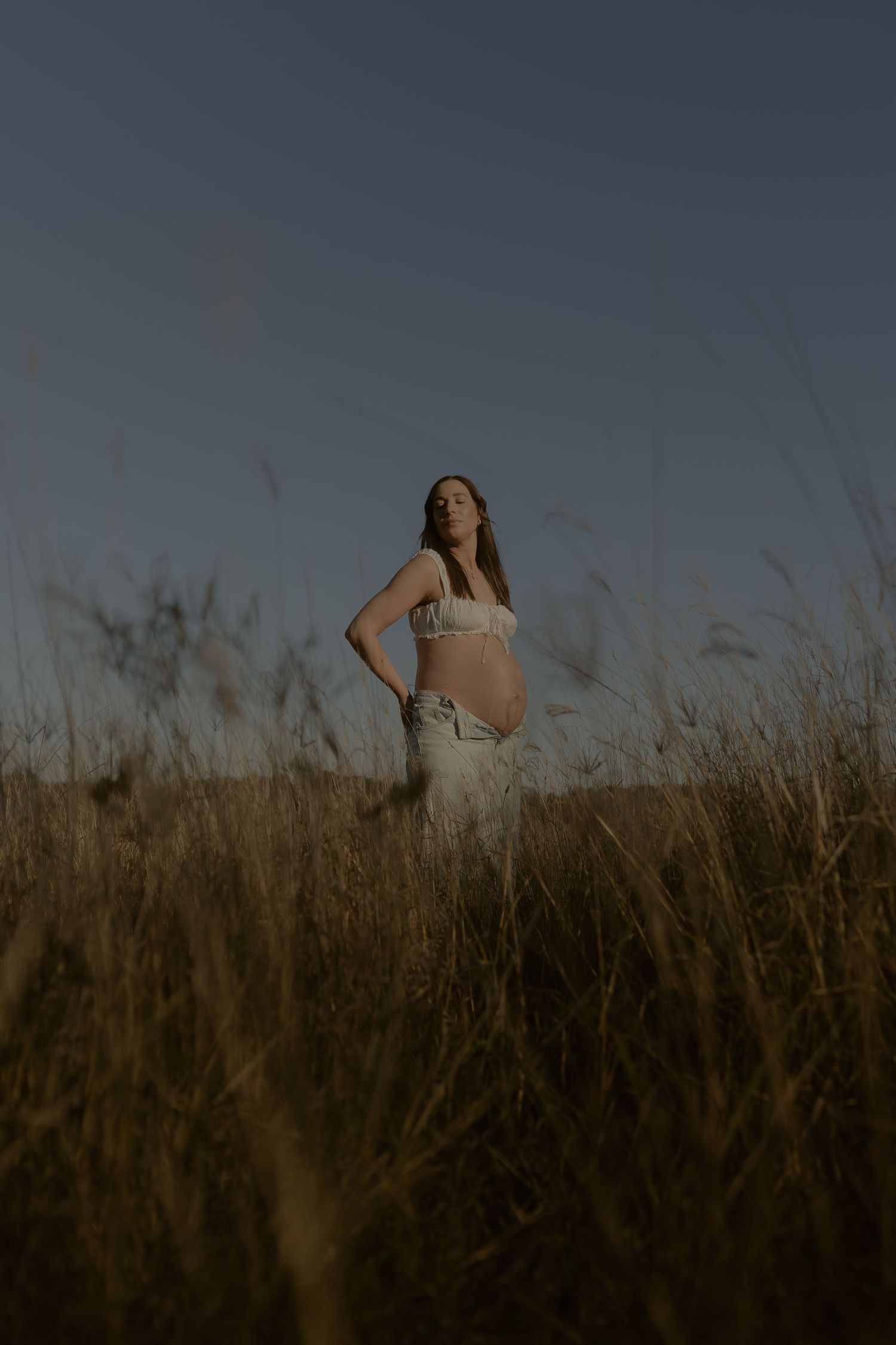 Pregnant woman in white crop top and jeans standing in tall golden grass field at dusk.