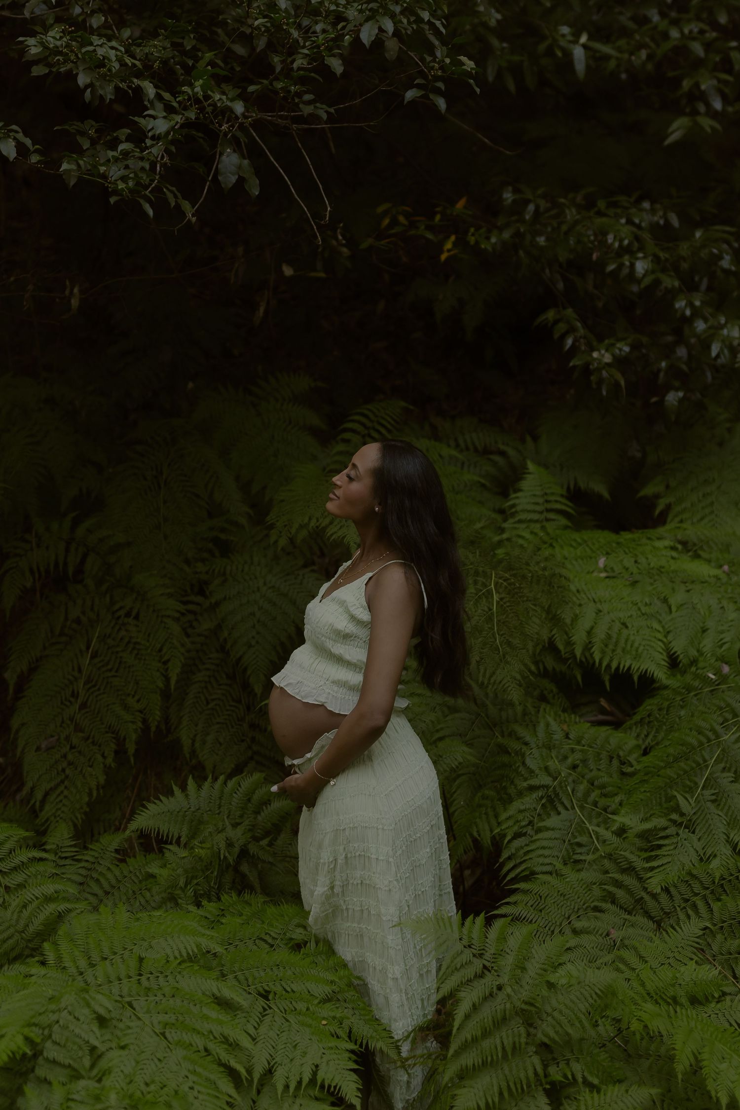 Pregnant woman in white dress cradling baby bump surrounded by lush green ferns in a forest.
