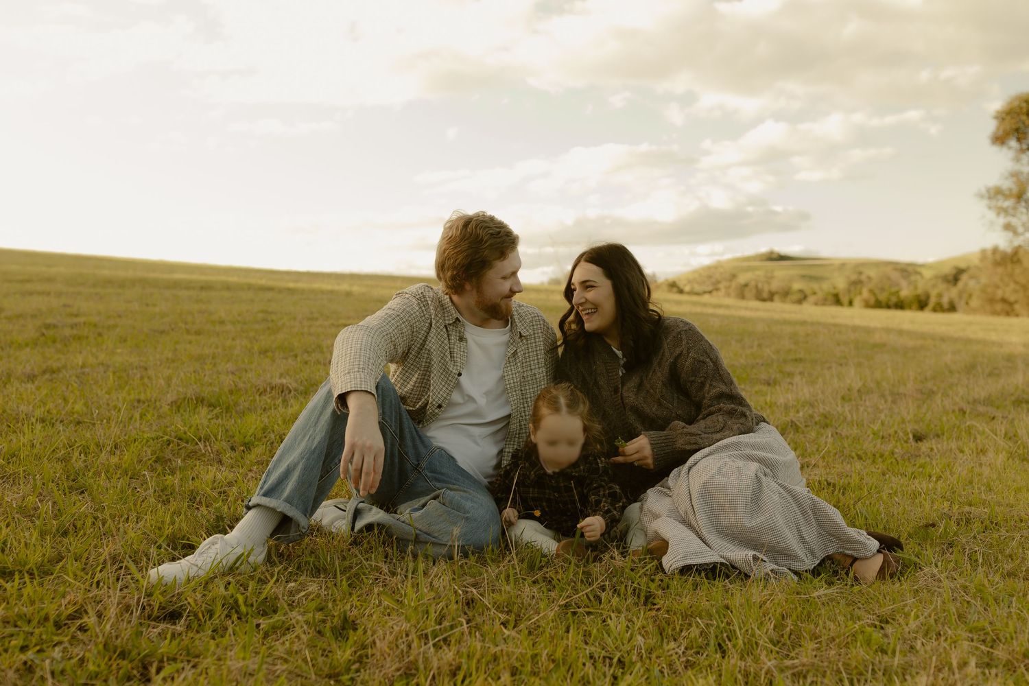 Happy family with toddler sitting together on green grass field at sunset, enjoying a peaceful outdoor moment.