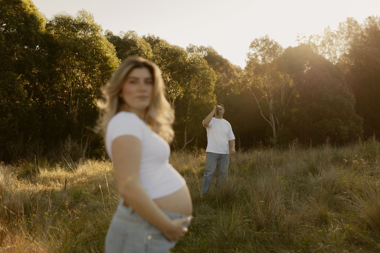 Pregnant woman in white top holding baby bump in golden sunlit field with partner walking away in background.