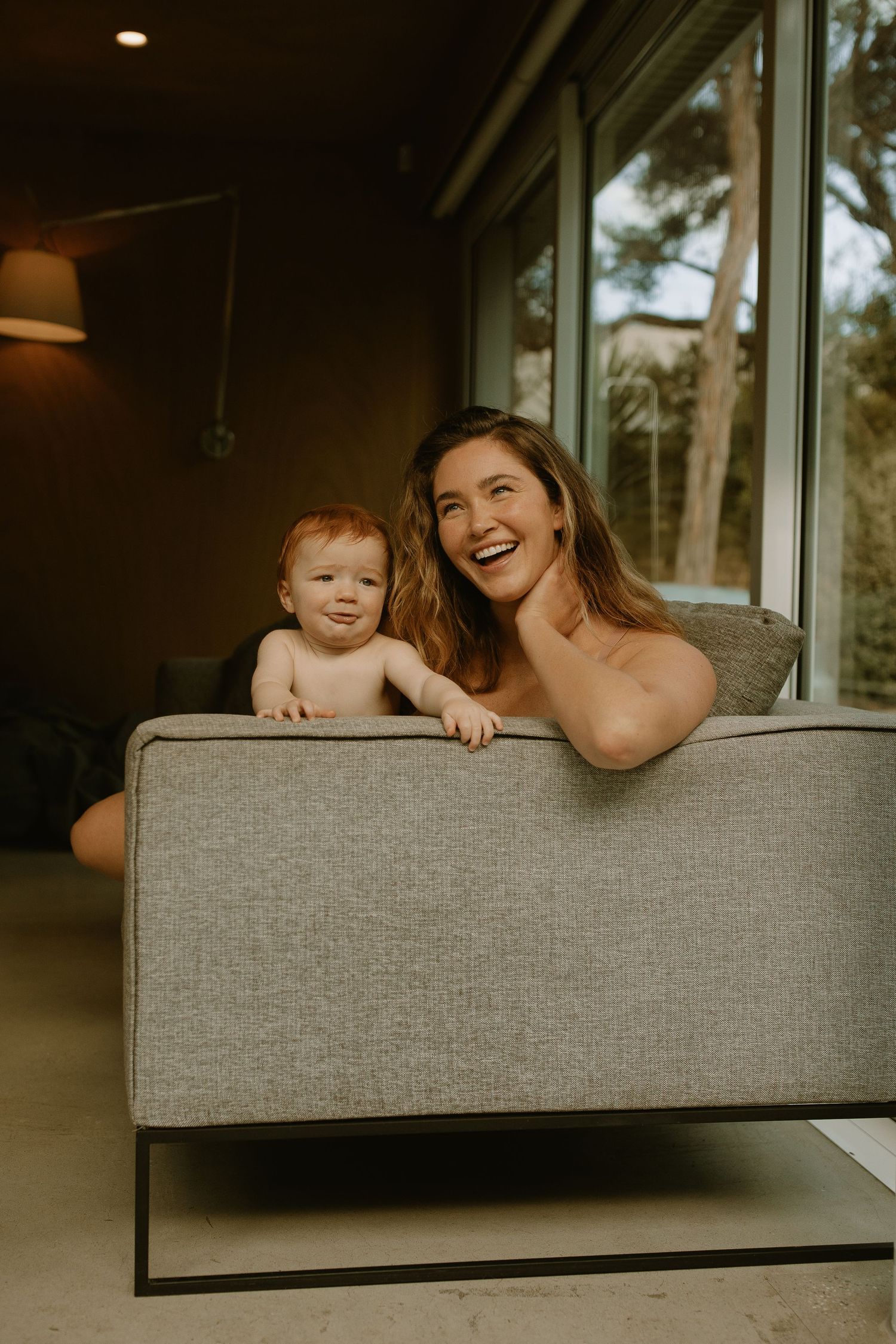 A warm indoor portrait of a parent and baby sharing a joyful moment while leaning over a modern gray couch.