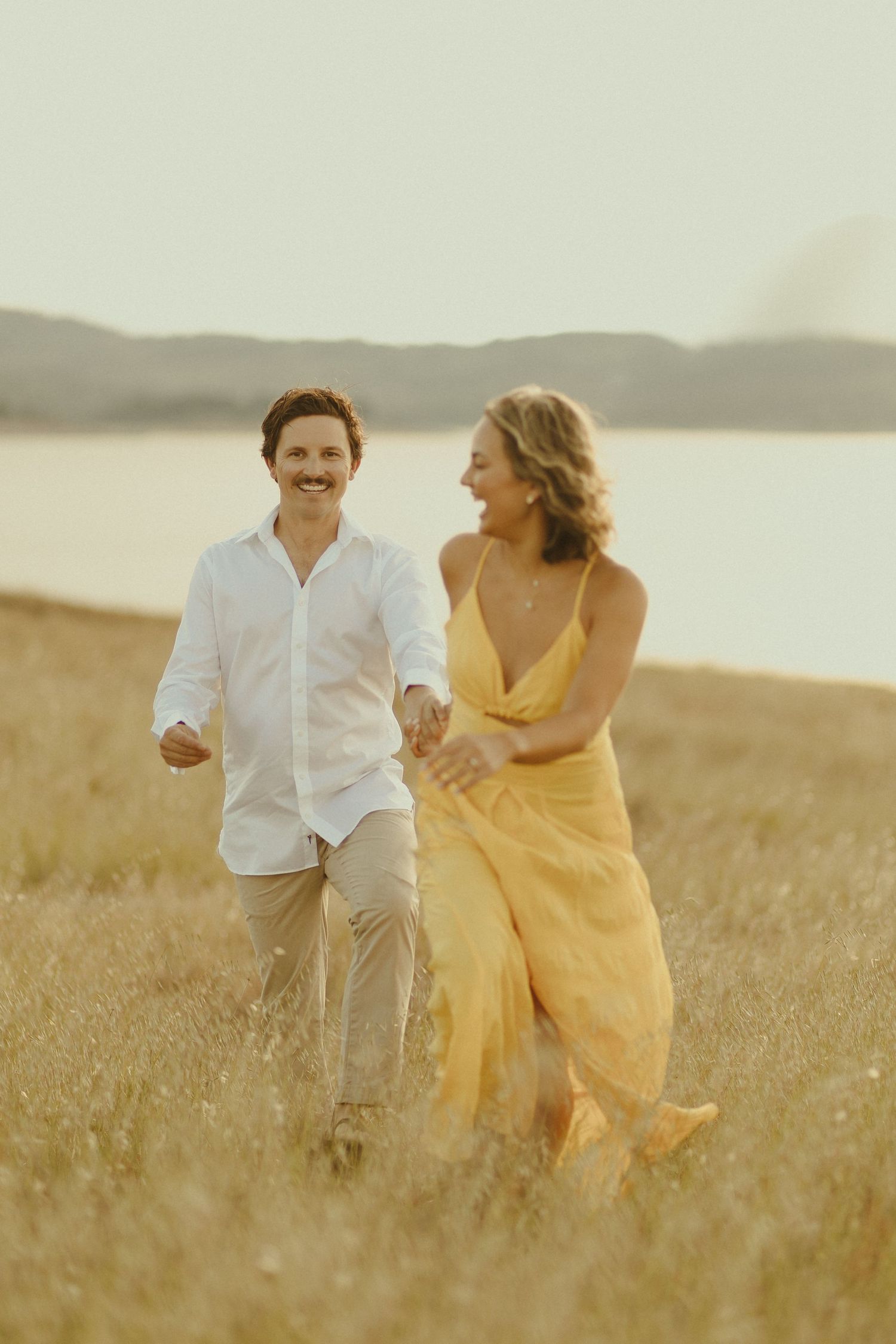 A couple runs together through a golden field at sunset with mountains and water in the background.