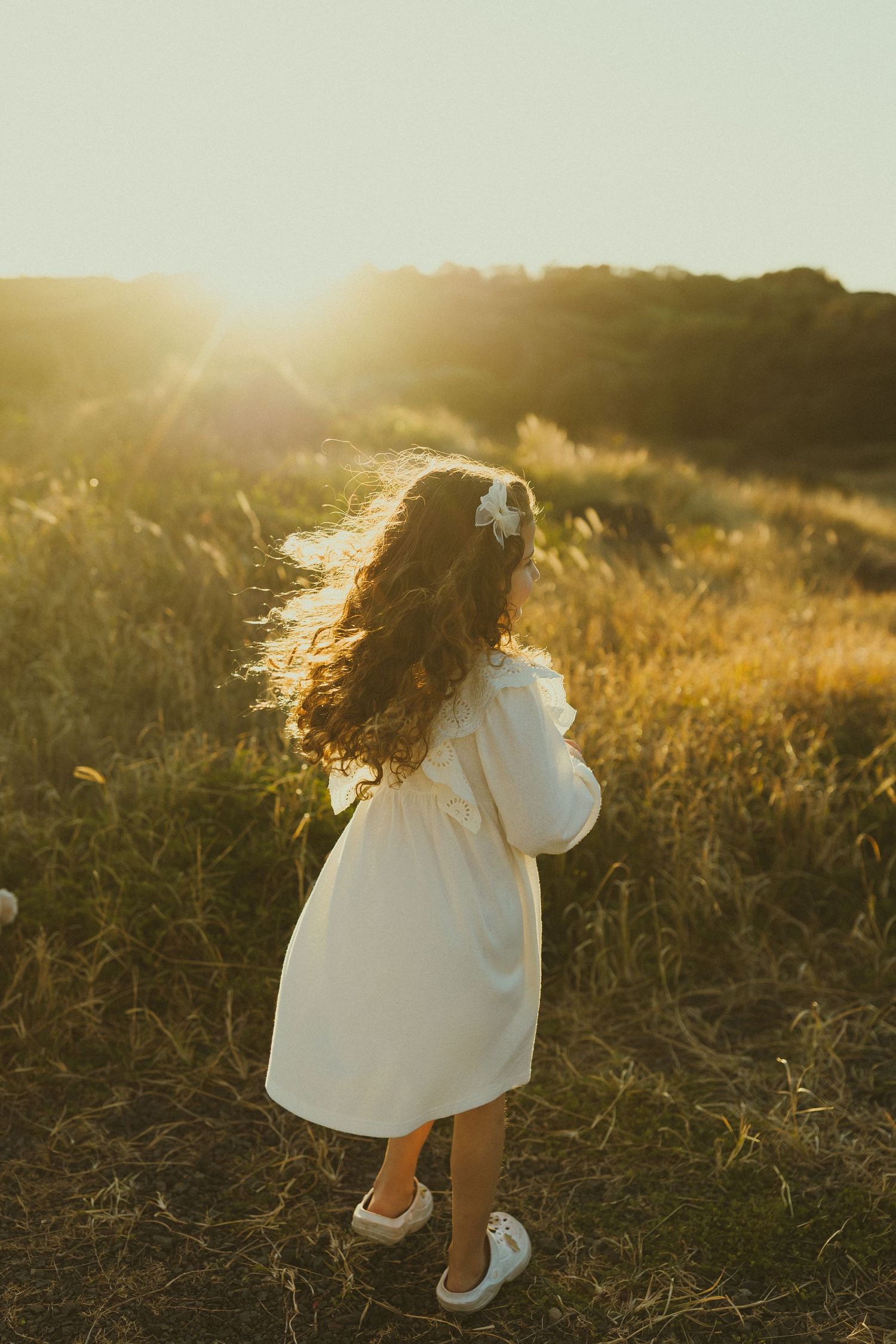 Figure in white dress twirls in sunlit field of golden grass during sunset.
