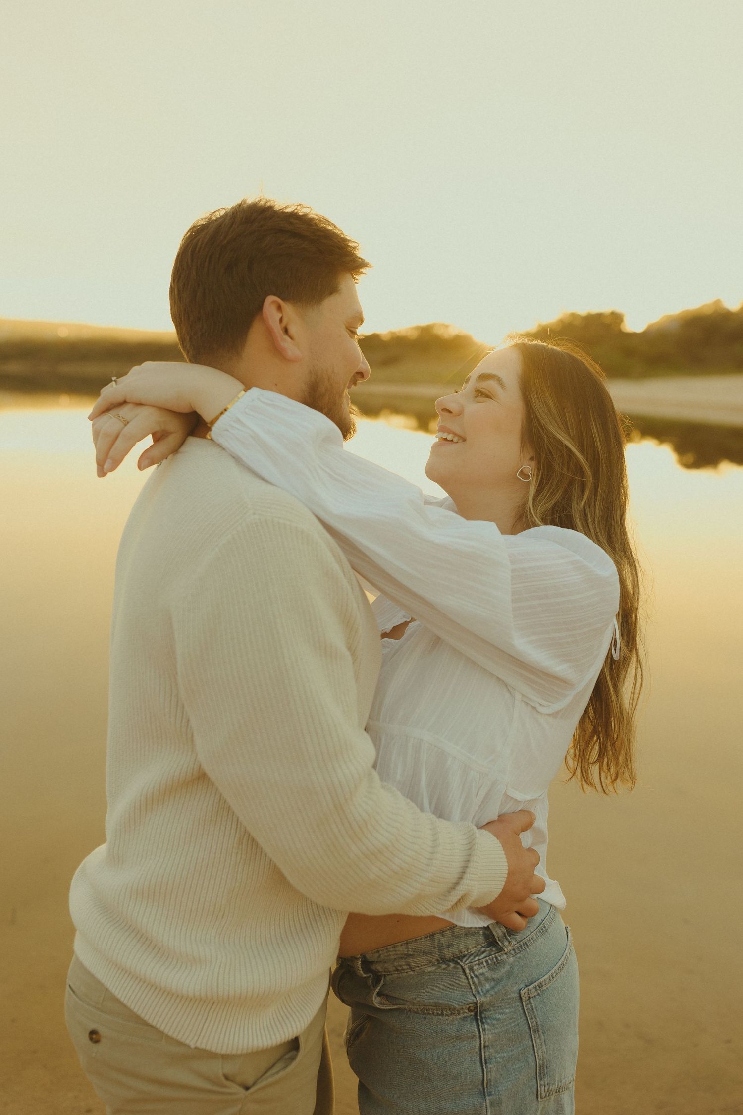 A romantic couple embraces while wearing cozy white sweaters during a warm sunset at the beach.