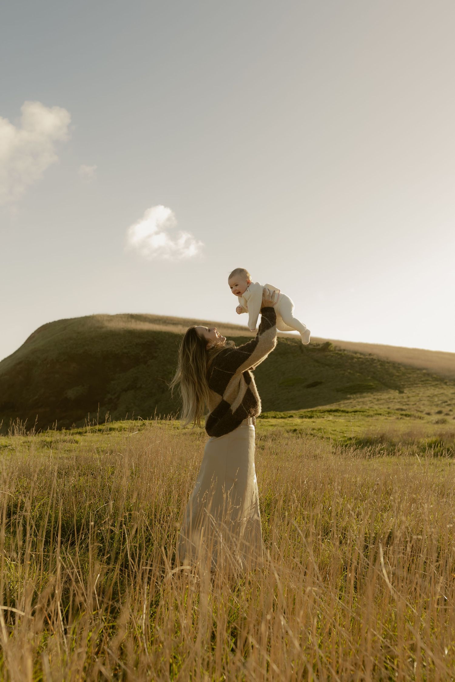 A mother joyfully lifts her baby in a sunlit green meadow with rolling hills in the background.