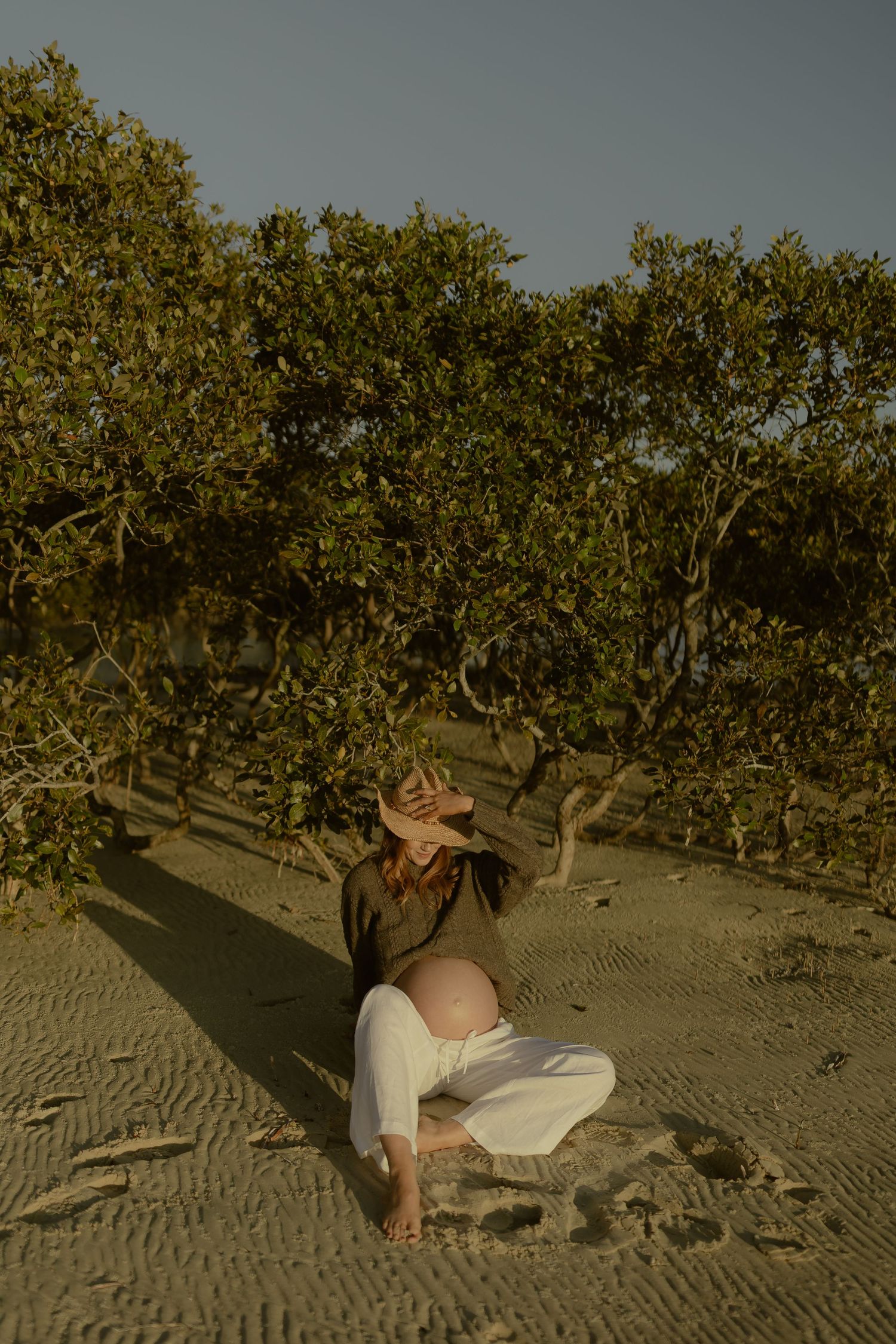 Pregnant woman in hat and white pants sits on sandy beach surrounded by lush green mangrove trees.
