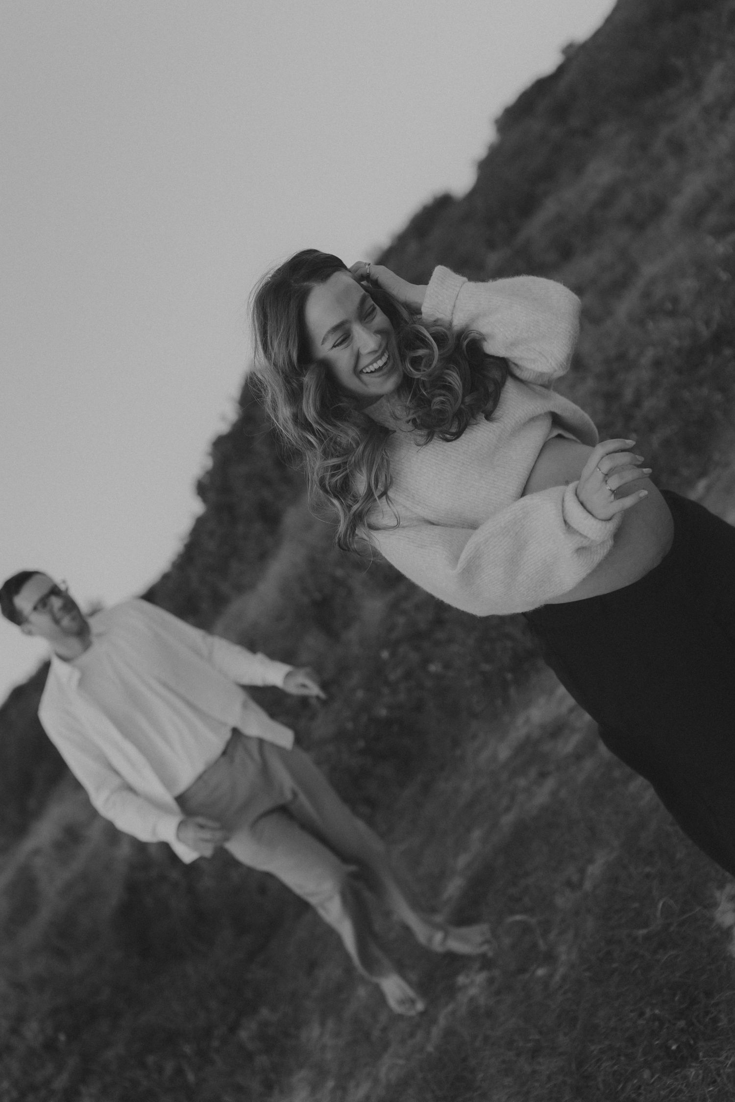 Black and white photo of laughing pregnant woman and man walking outdoors on hillside.