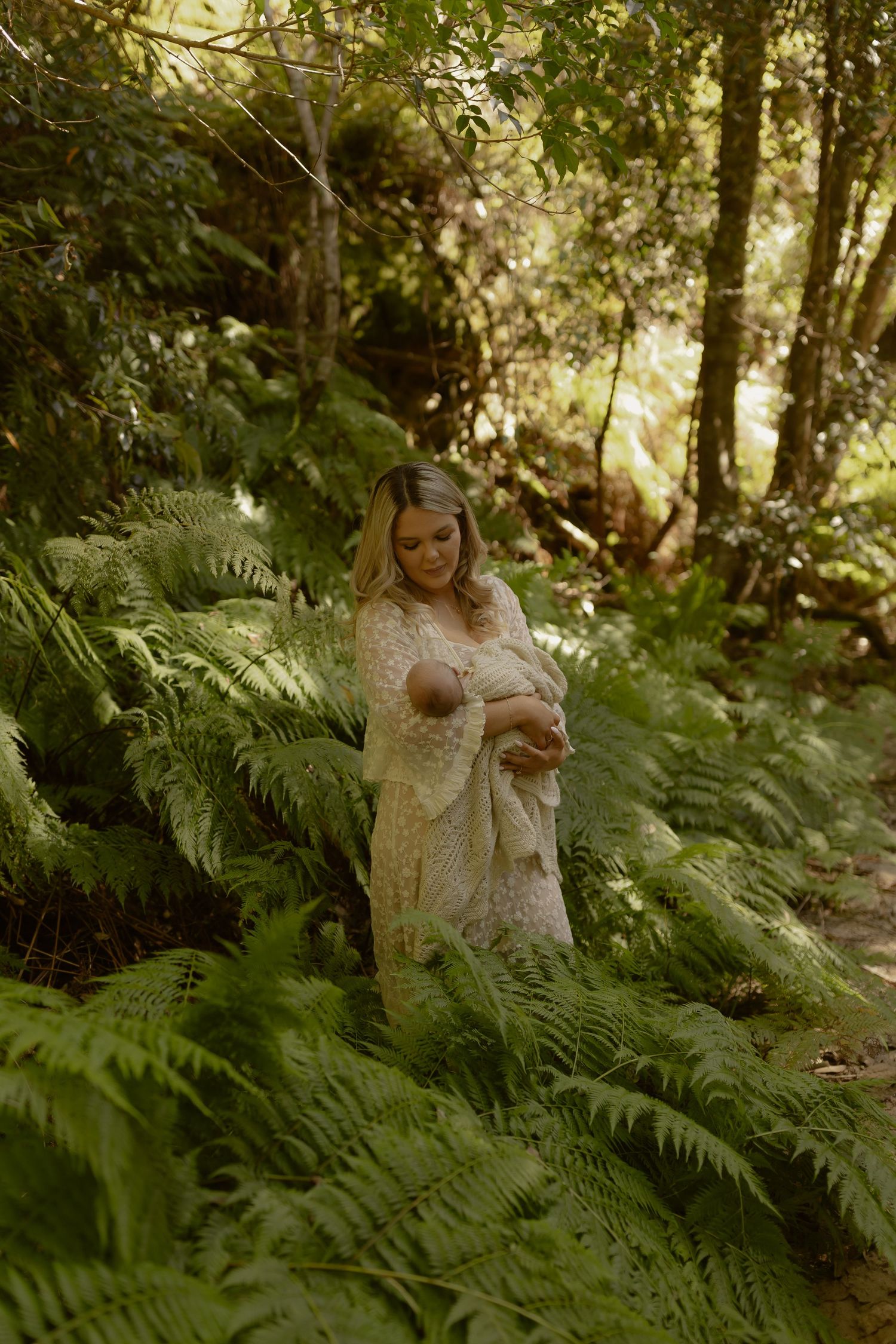 A mother in a lace gown cradles her newborn among lush green ferns in a serene forest setting.
