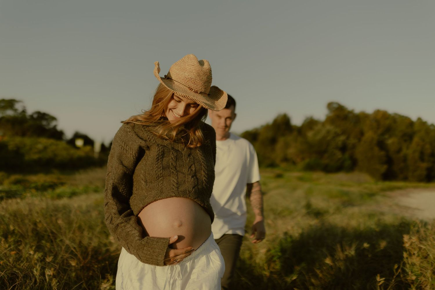 Pregnant woman in cowboy hat and knit sweater stands with partner in golden hour field.
