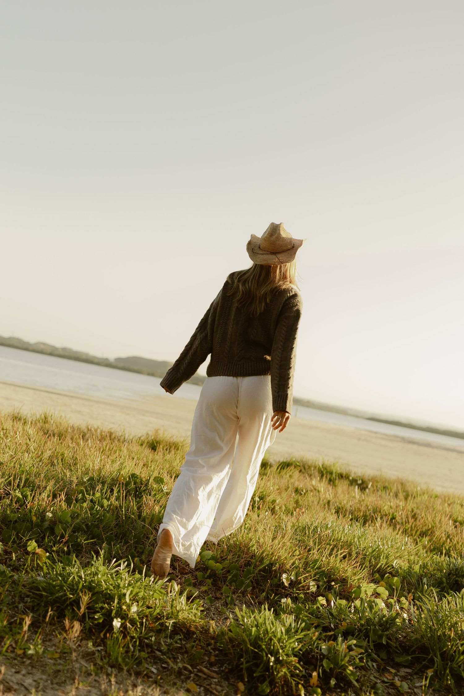 Woman in white pants, dark jacket, and straw cowboy hat walking through coastal grass toward the water at sunset.