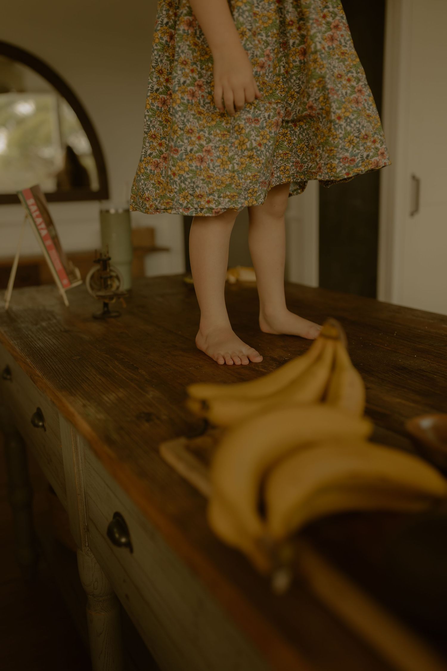 Young barefoot girl in floral dress standing on rustic wooden table beside fresh yellow bananas.