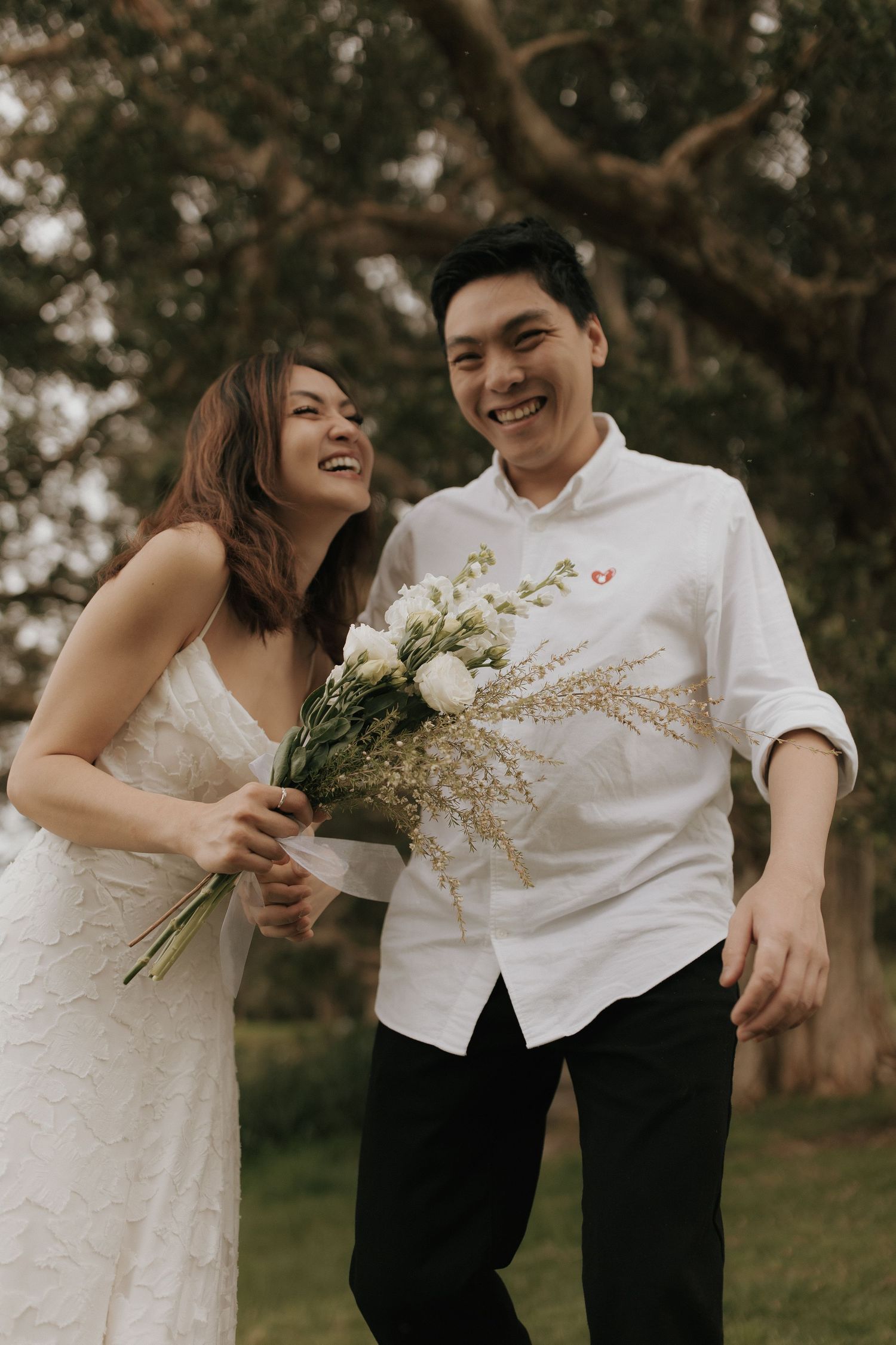 A couple in wedding attire shares a joyful moment while holding dried flowers in an outdoor garden setting.