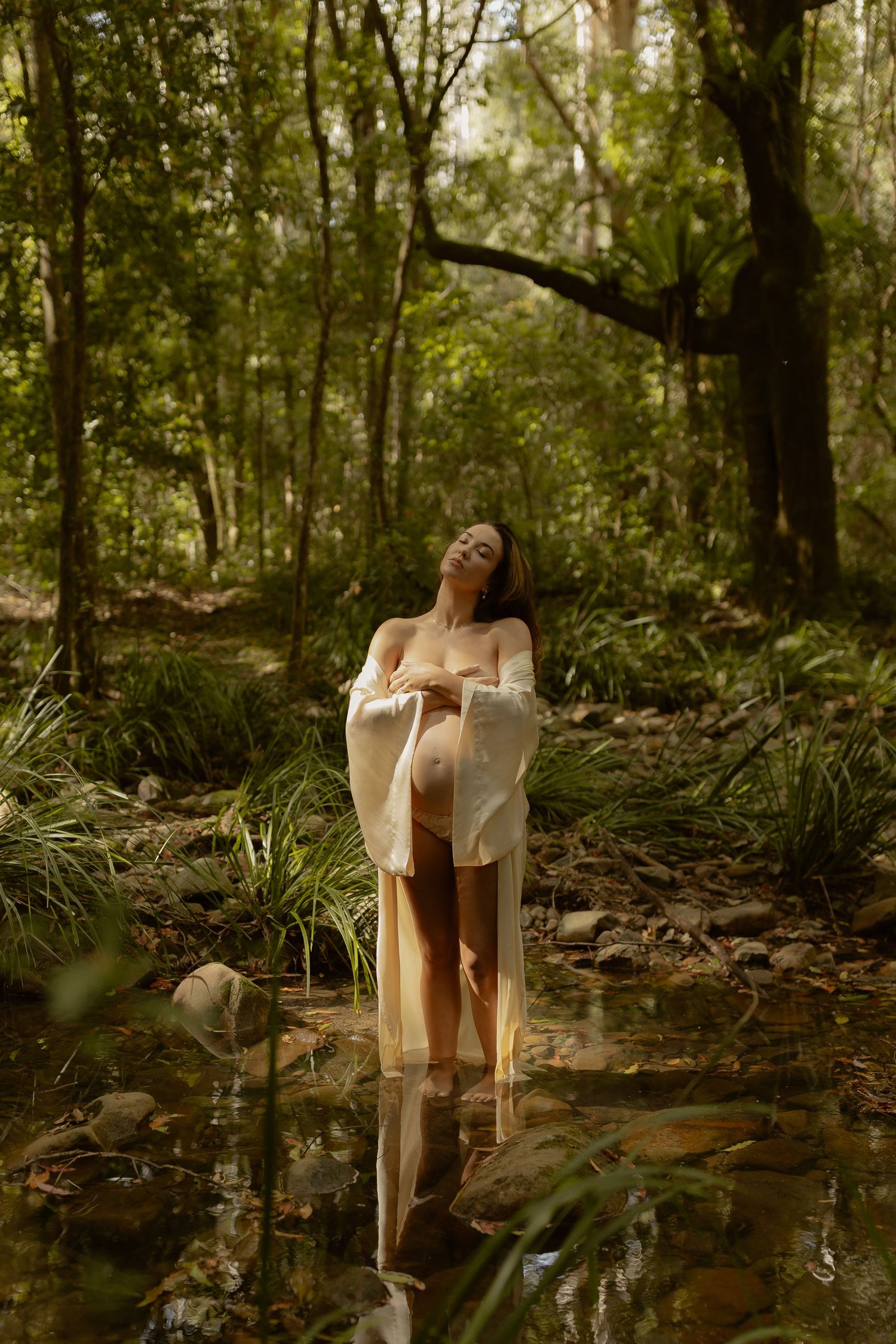 Artistic portrait in a sunlit forest stream with flowing white fabric creating an ethereal mood.