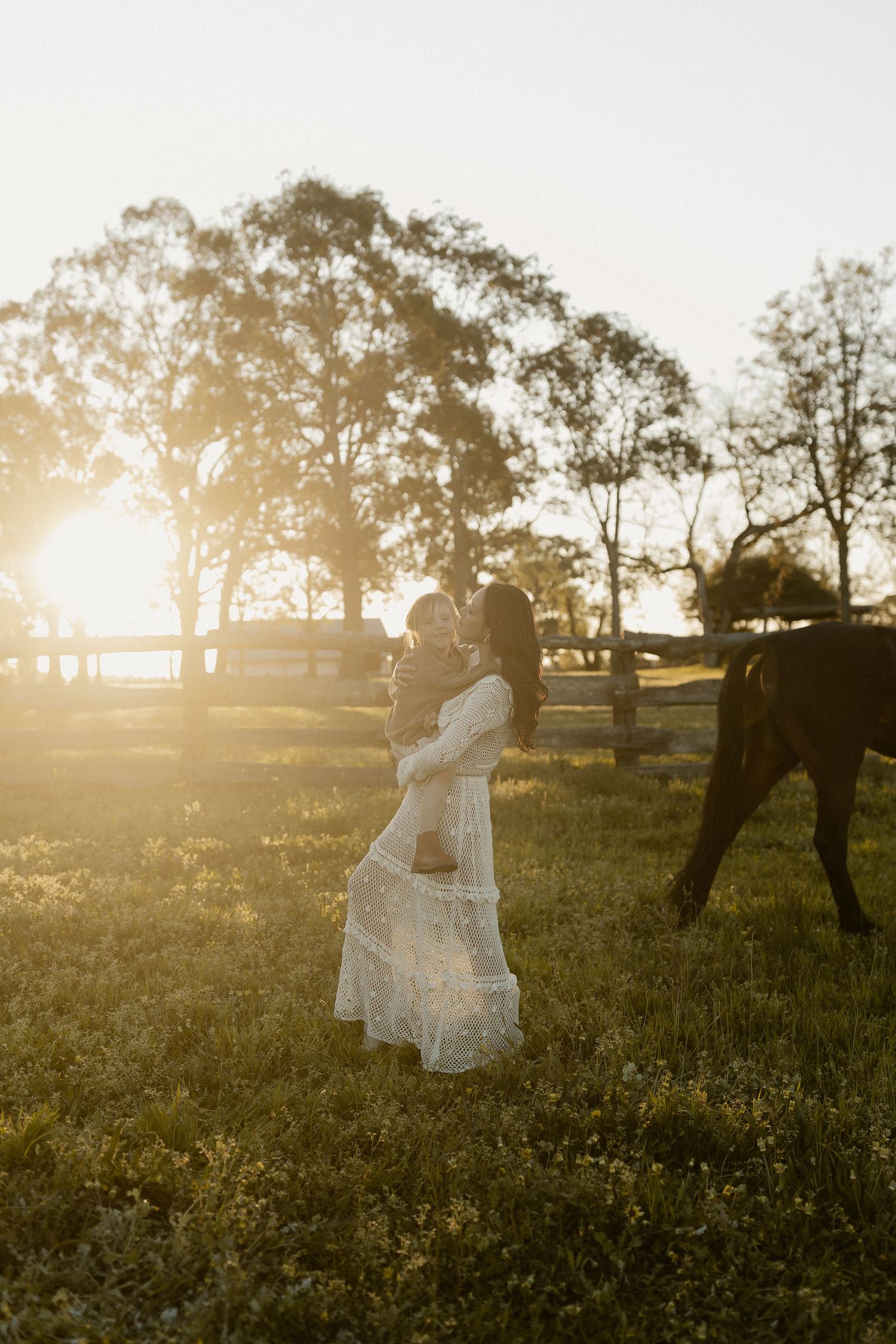 Silhouetted in golden sunset light, someone in a flowing white dress stands in a grassy field as a horse grazes nearby.