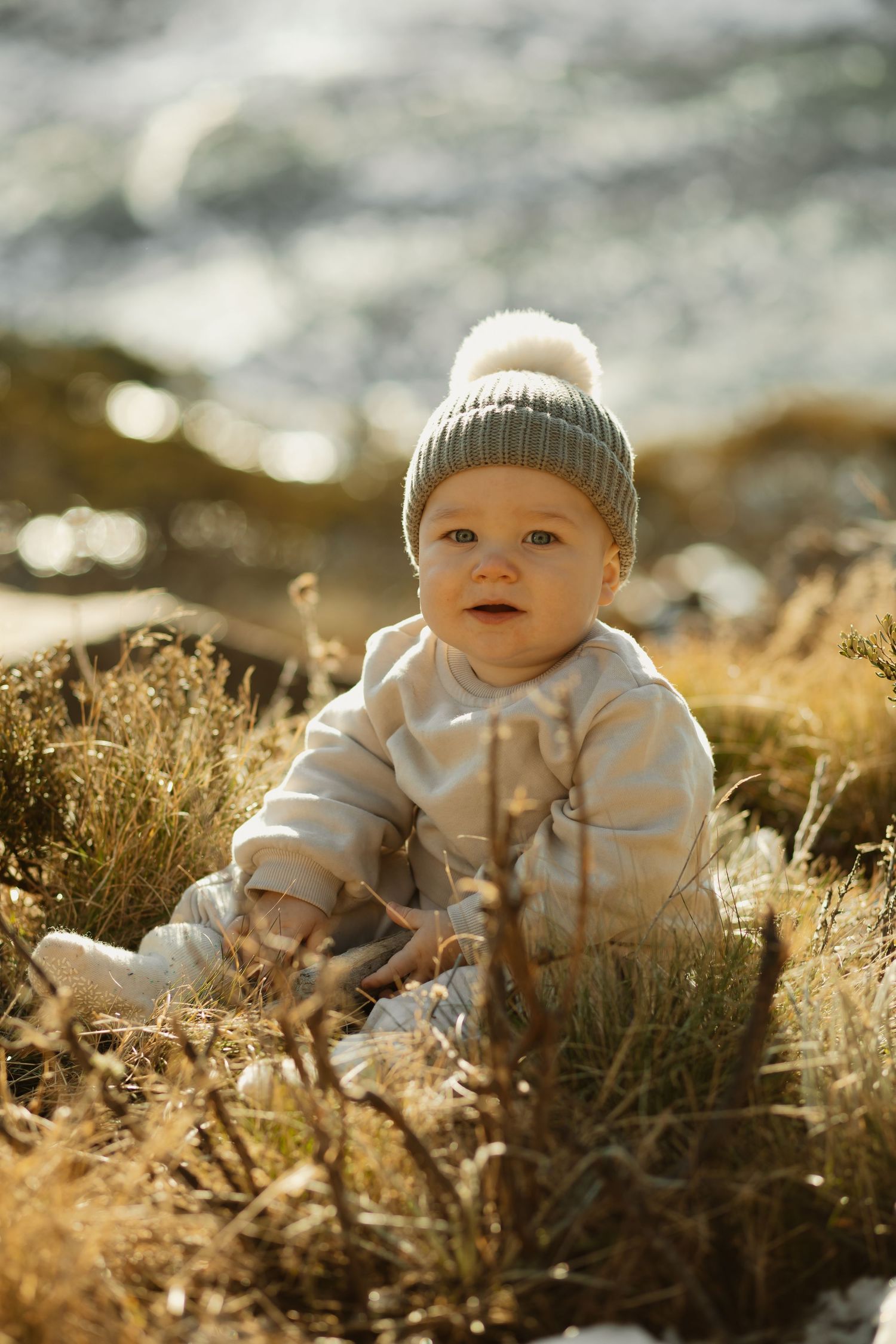 Small child wearing a winter hat and white outfit sits among golden grass in sunlit outdoor setting.
